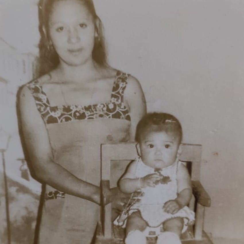 Foto em plano aberto antiga em preto e branco de uma mulher jovem, de cabelos longos semi-presos e vestido de alça, ao lado de uma cadeirão de madeira com uma bebê de vestido, sentada.