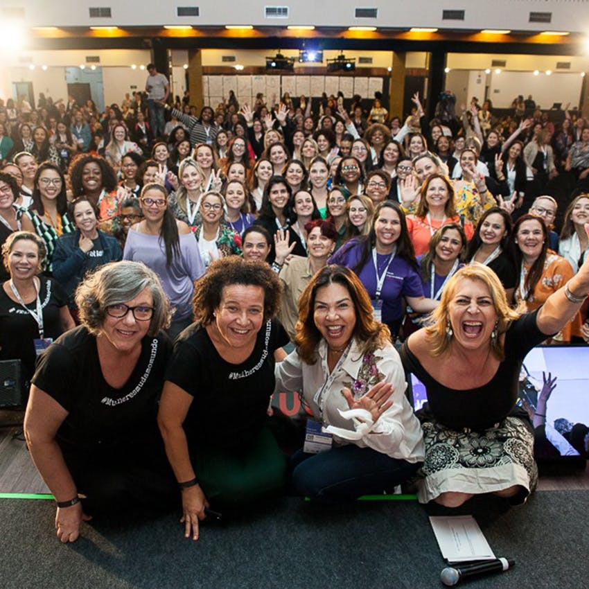 Foto em plano aberto de uma grande sala com dezenas de mulheres de diversas idades, sorrindo e olhando na direção da câmera em um grande evento.
