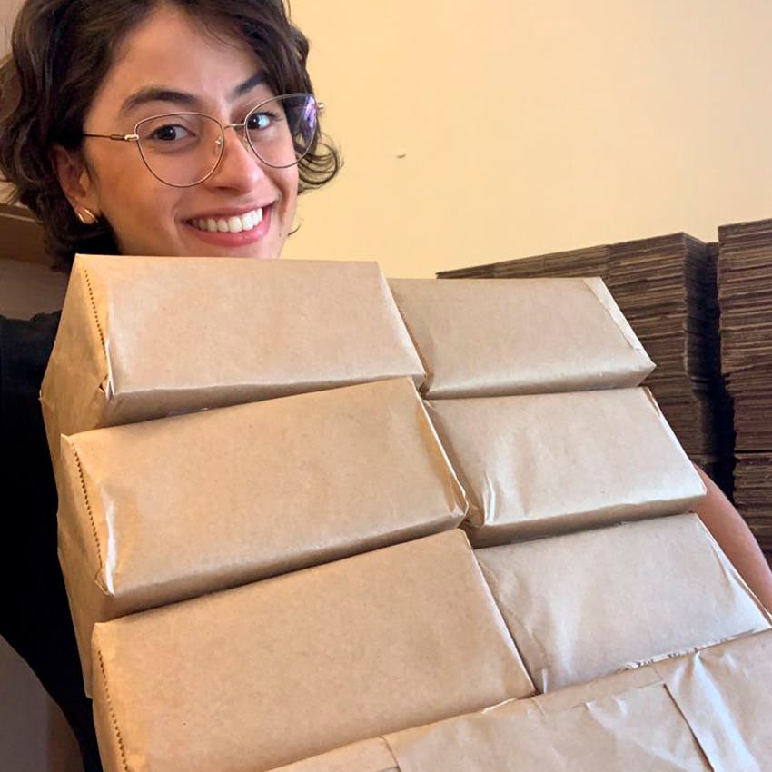 Foto em plano fechado de uma jovem mulher, de cabelos curtos e castanhos, óculos, sorrindo. Ela segura sete caixas embrulhadas em papel pardo.