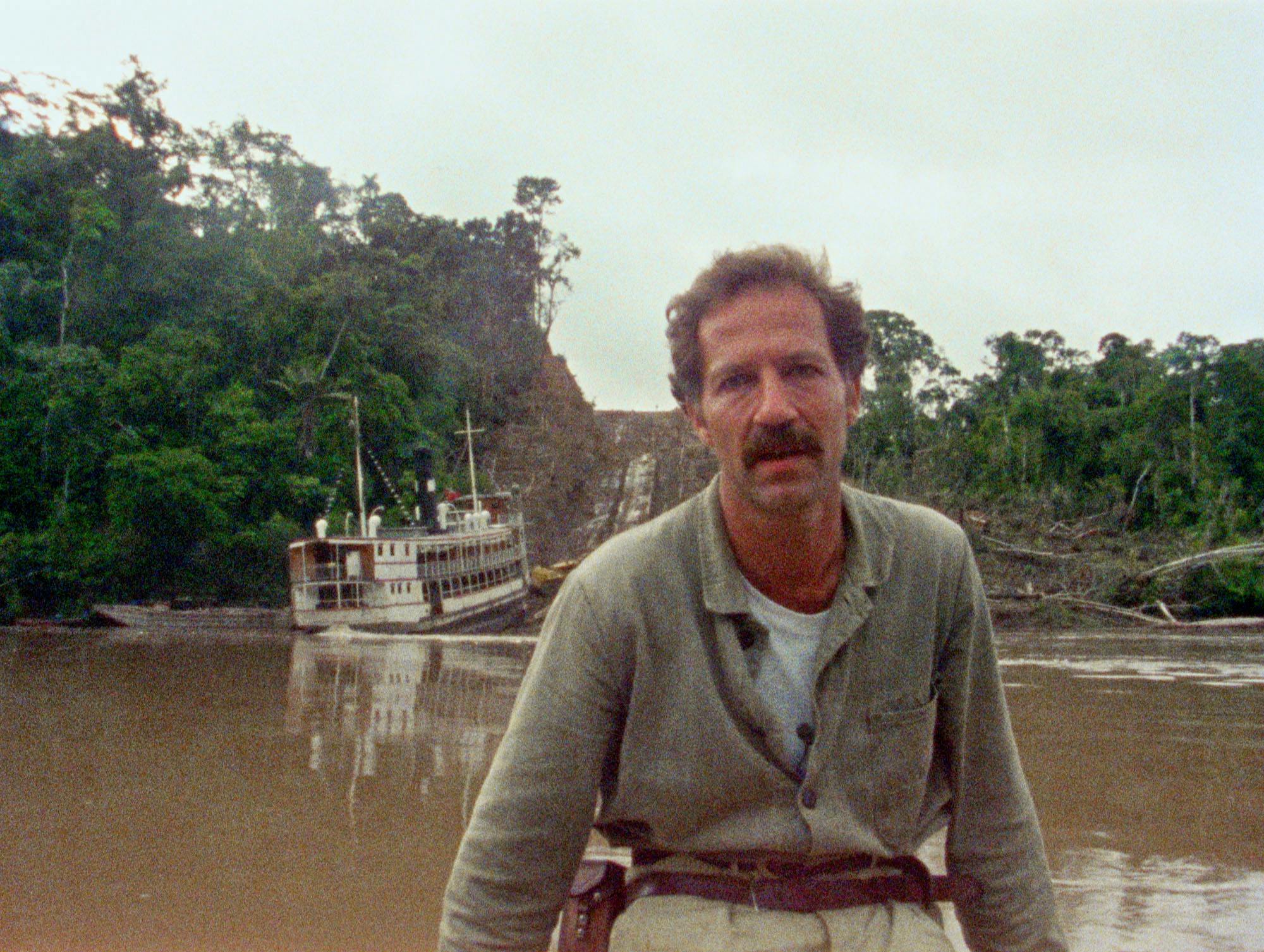 Man sits by a river with boat in background.