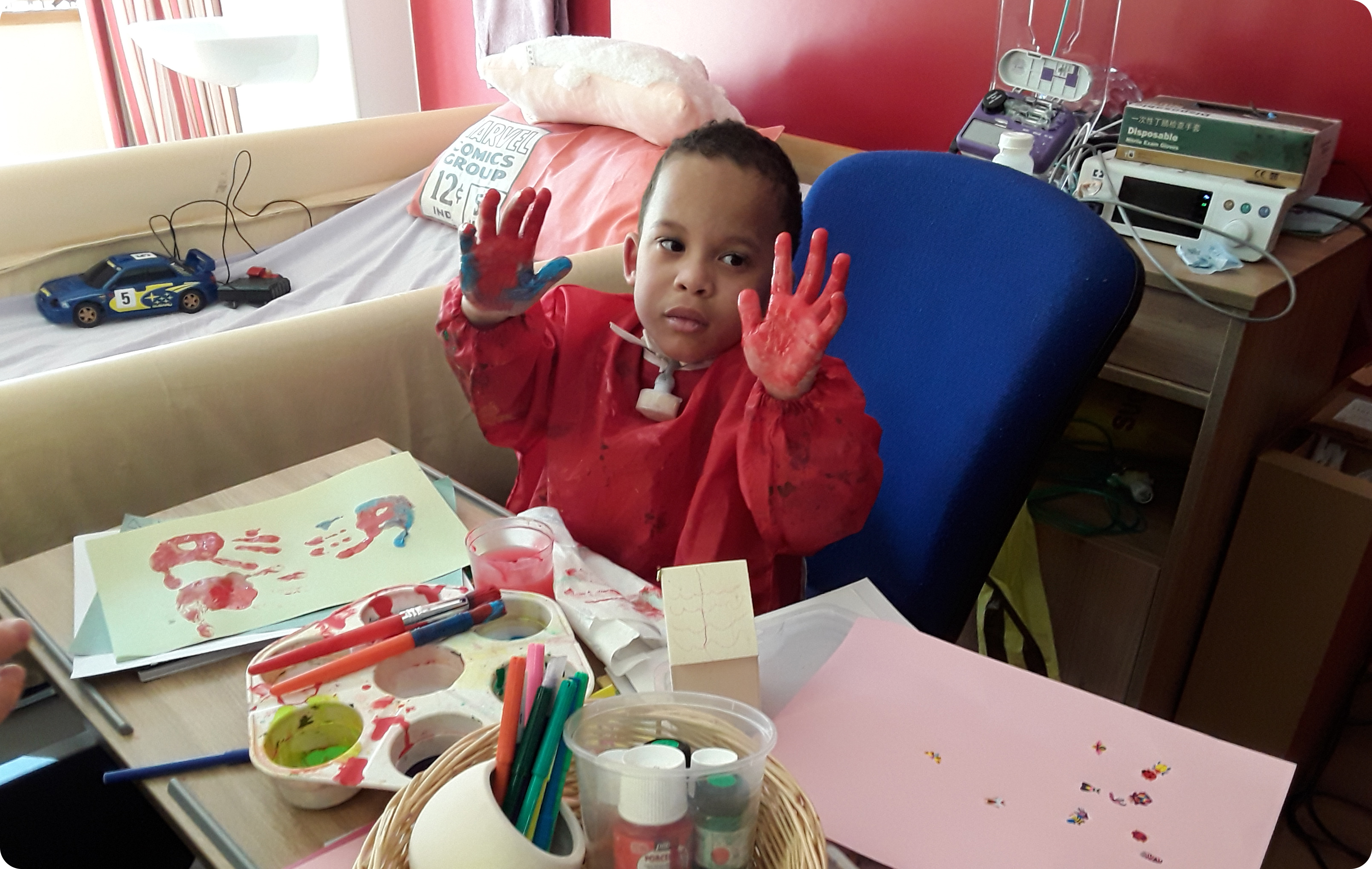 Picture of a young child painting, they are looking at the camera with their painted hands up
