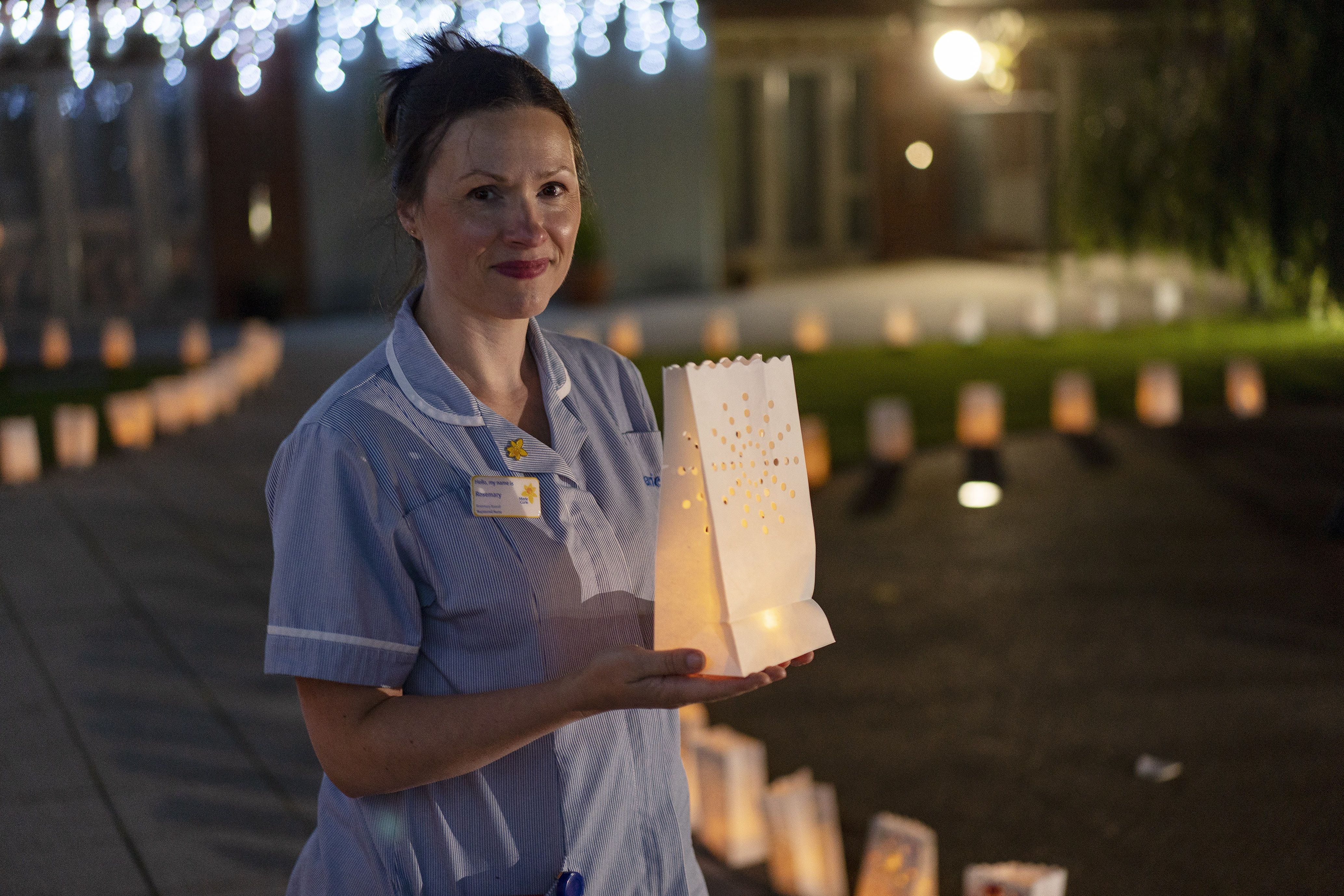 A woman in a nurses outfit holding a lantern, with a row of lanterns behind her lighting a pathway.