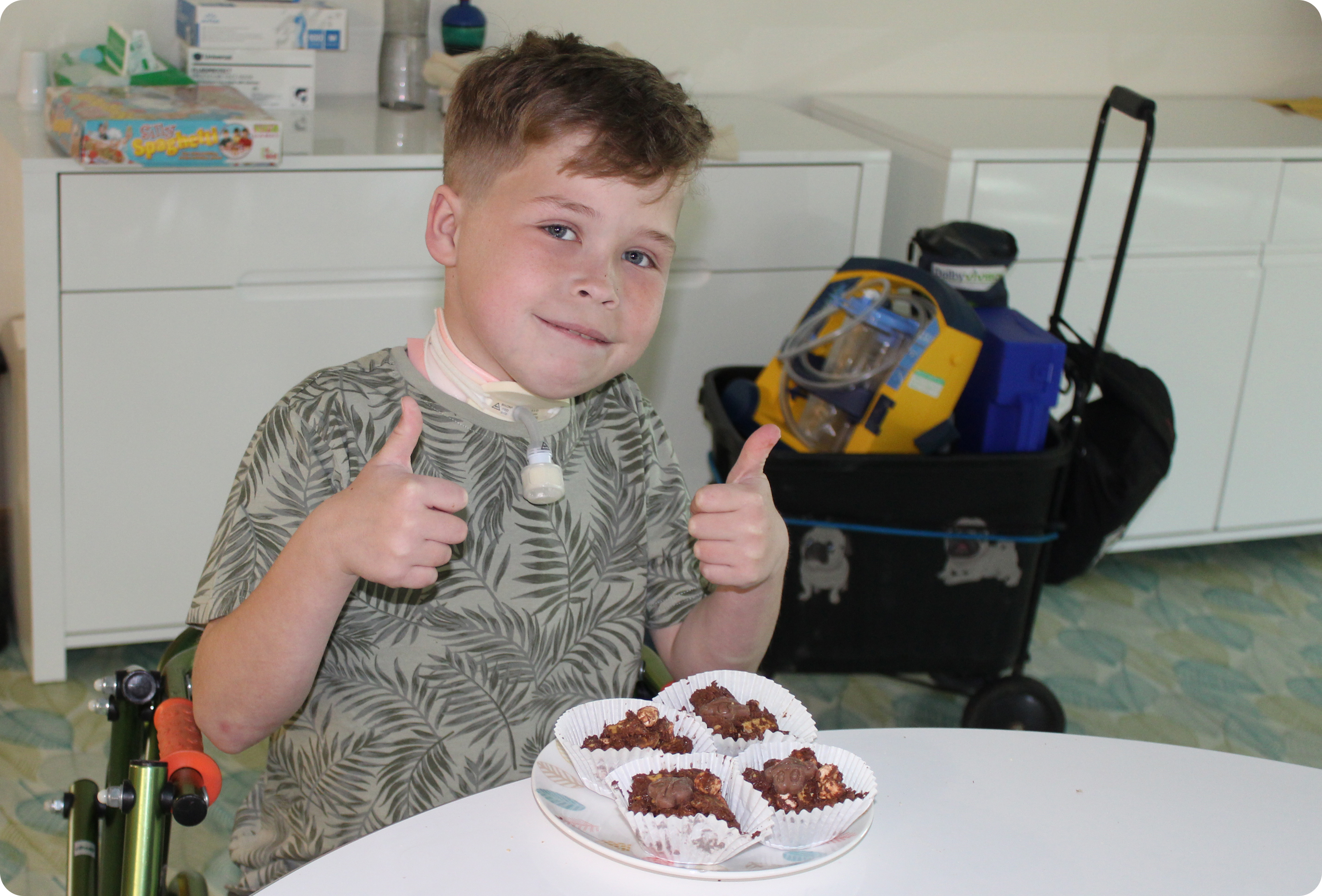 Picture of a child smiling towards the camera with their thumbs up and cakes in front of them