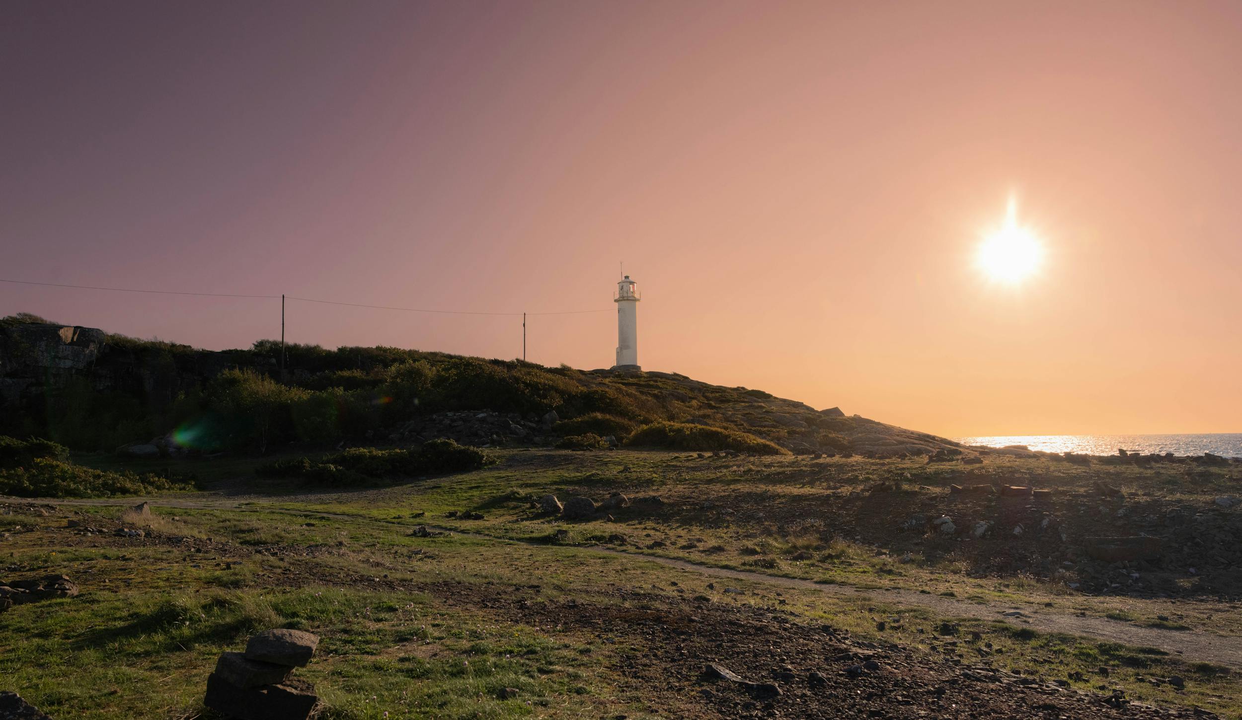Sonnenuntergang am Meer mit einem Leuchtturm auf einem Hügel.