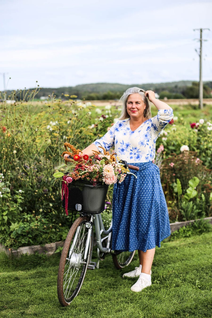 Hanna Wendelbo står framför en blomsteräng med cykelkorgen full av blommor.