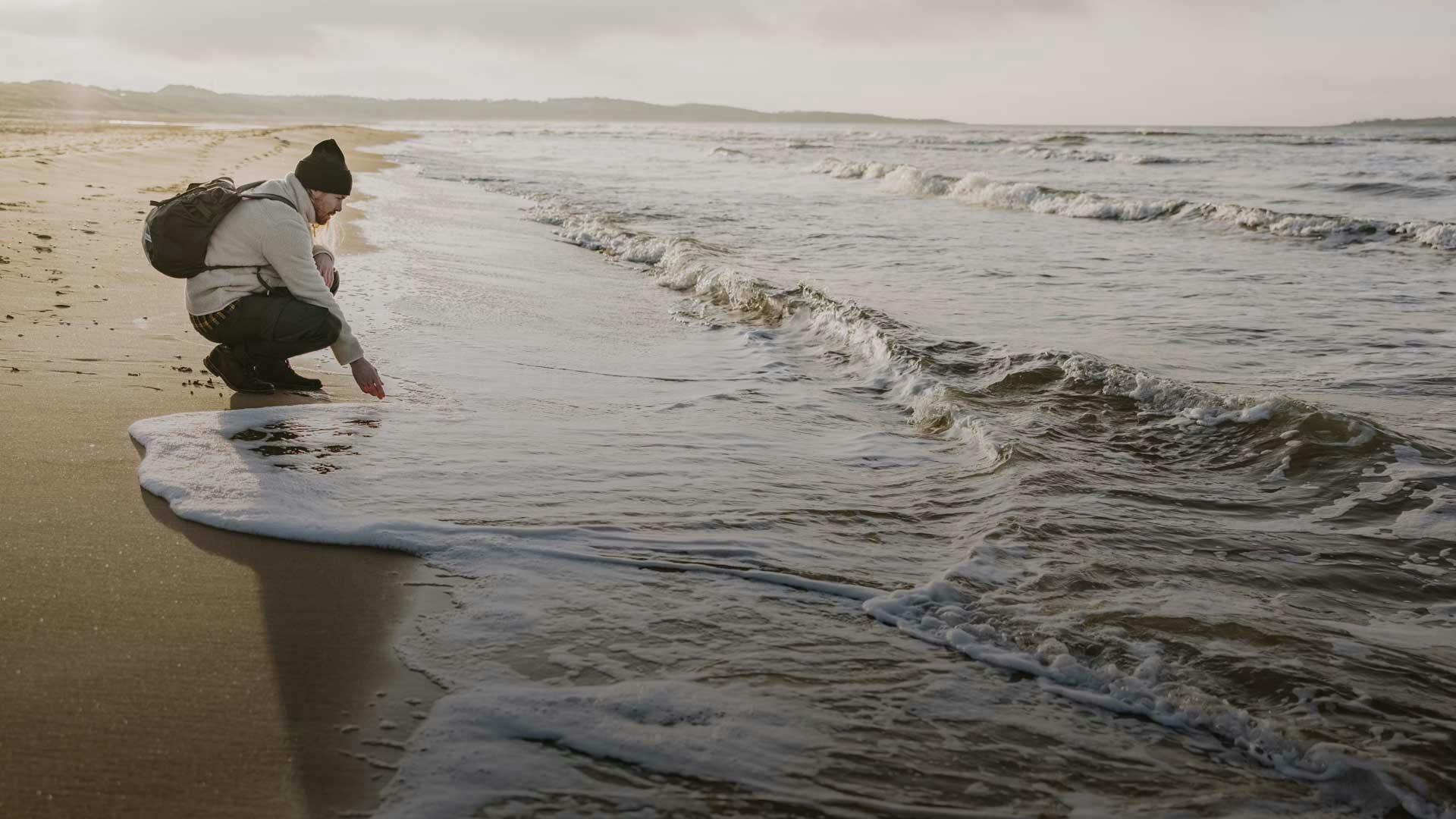 Mann mit Rucksack kniet am Strand im Herbst und hält Hand ins Wasser.