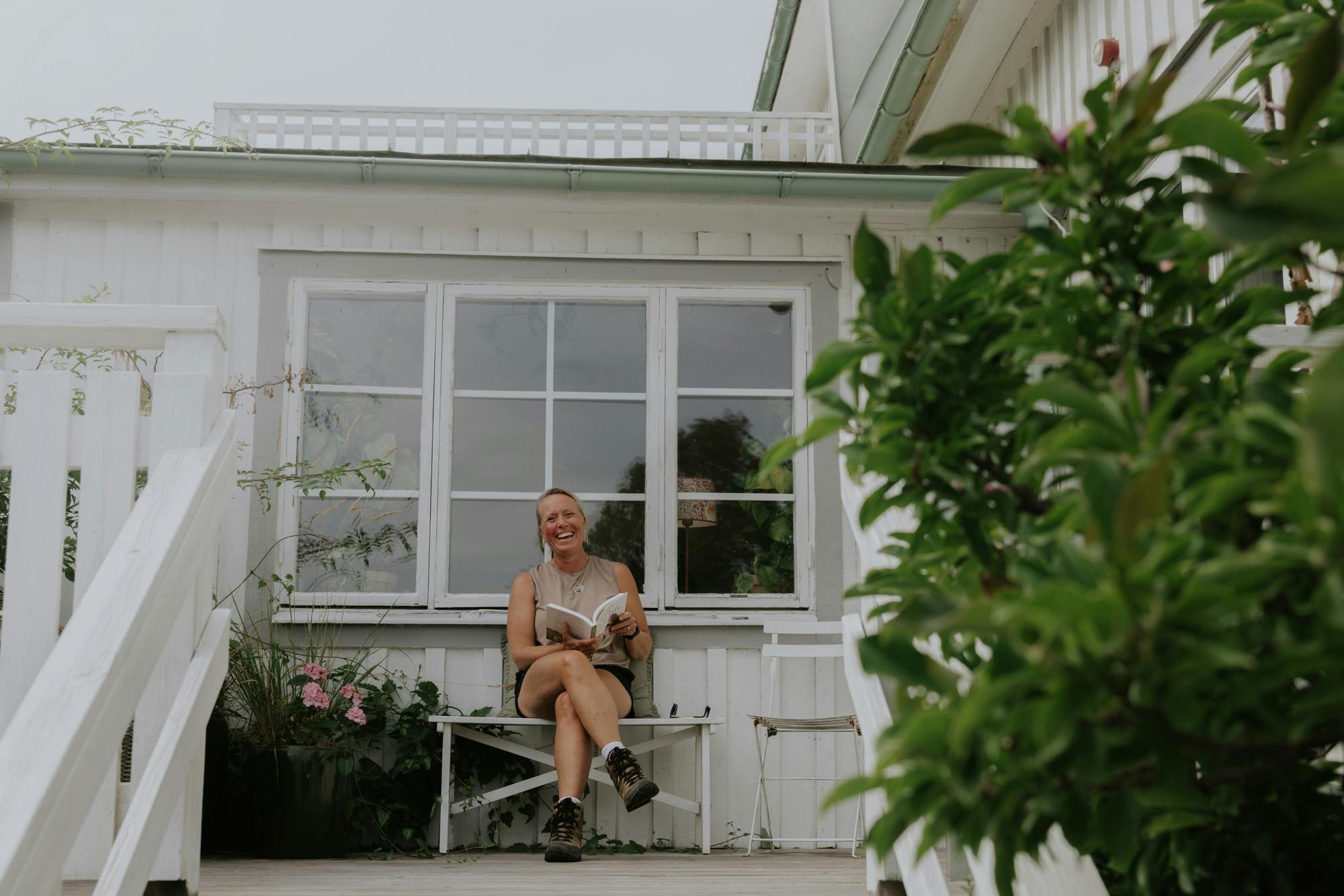 Steninge Kuststation, kvinna sitter på altanen och läser bok. Tittar in i kameran och ler.