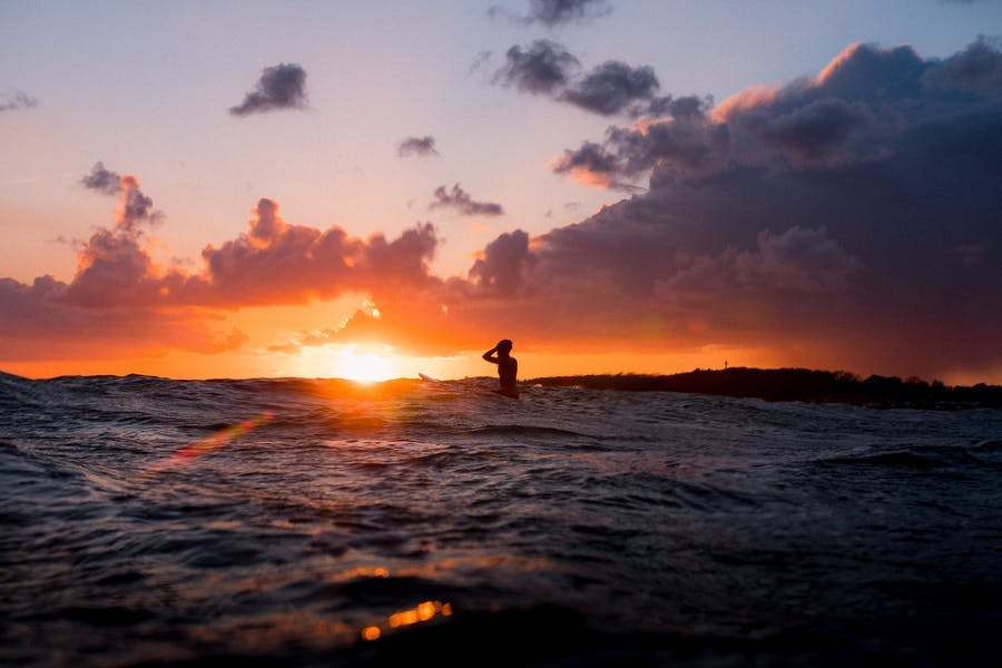 En person sitter på surfbrädan och blickar mot solen som är påväg ner i havet.