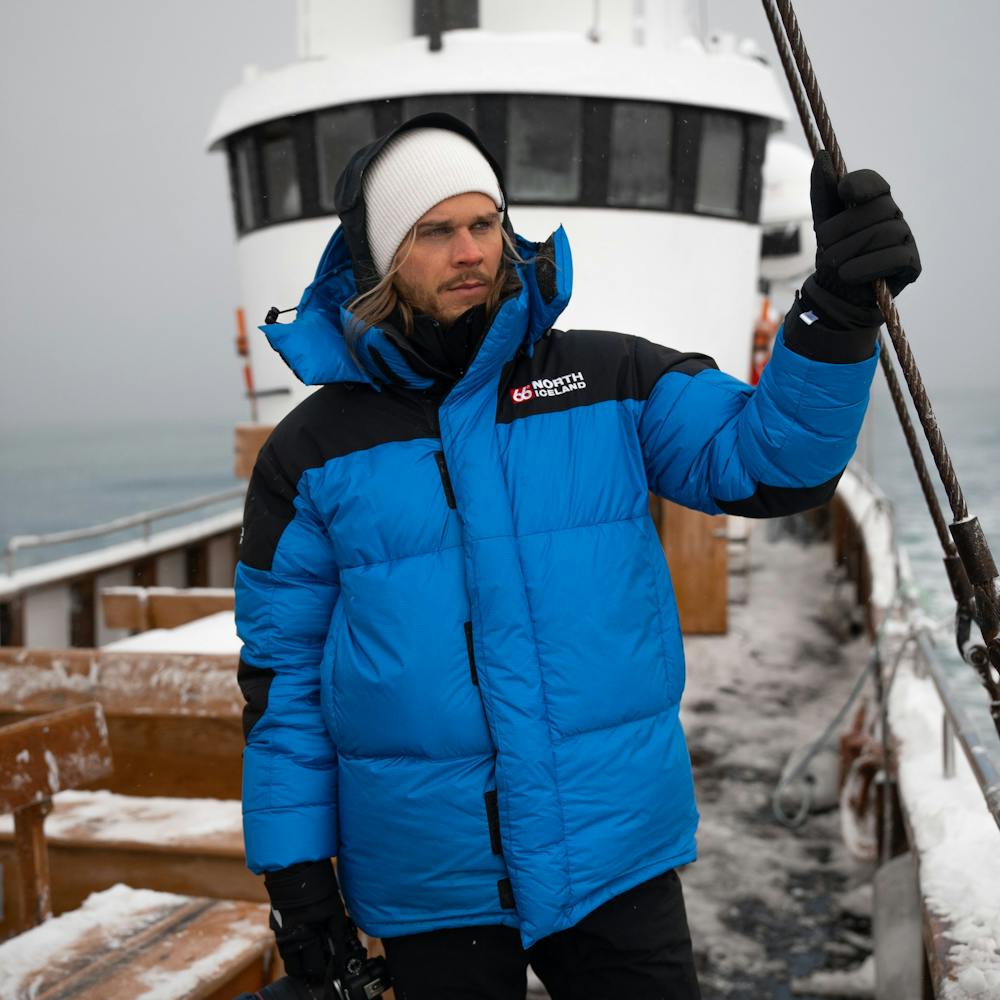 The Icelandic male model Rúrik Gíslason standing on a boat with snow on it. He wears a lightblue downjacket , a white beanie and thick gloves
