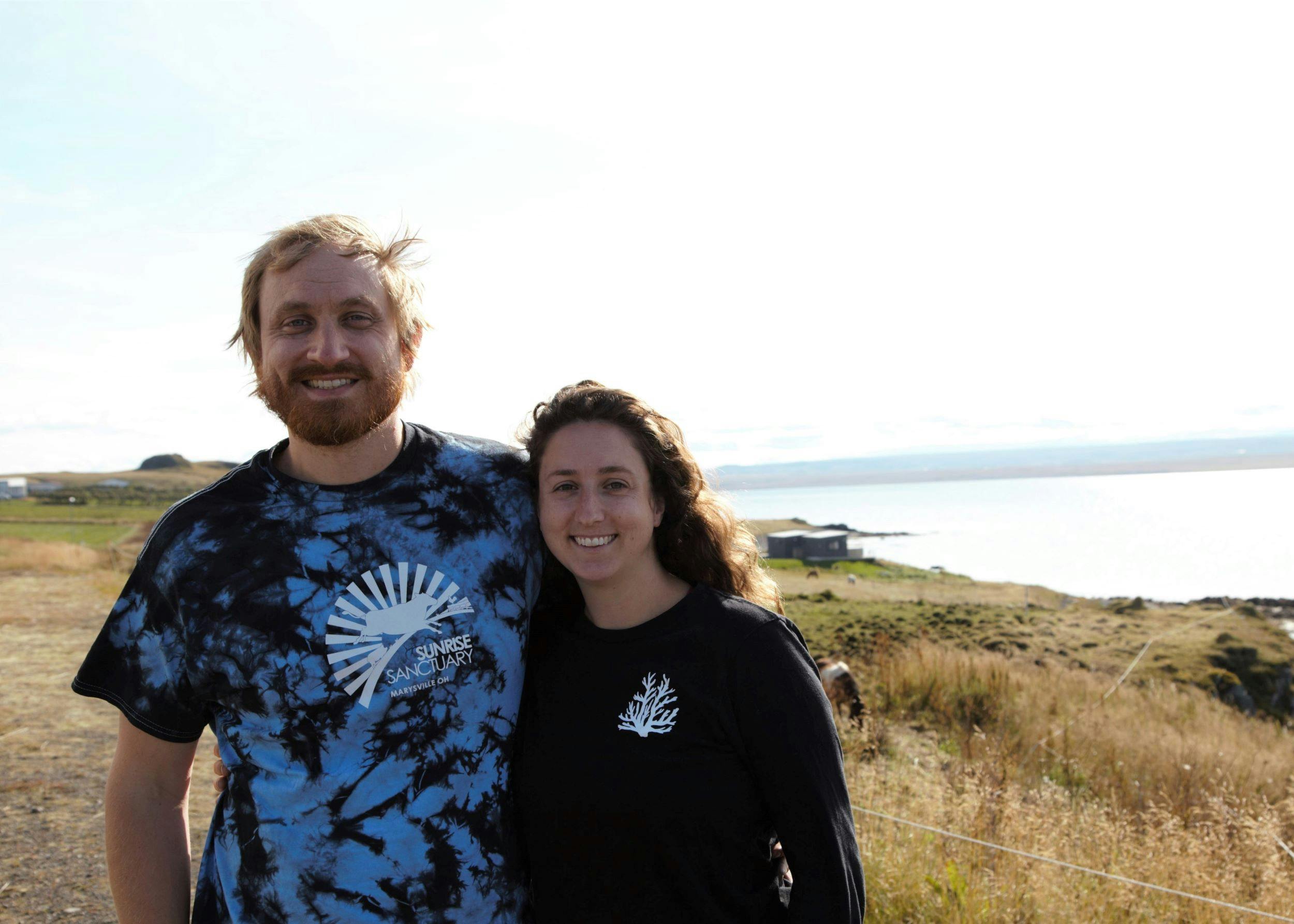 Young woman and man standing in front of a horse paddock, the ocean in the background