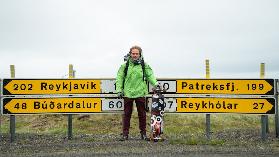 A young man in a green rain jacket and with a longboard in his hands stands in front of four roadsigns that show the distance to four different towns in Iceland.