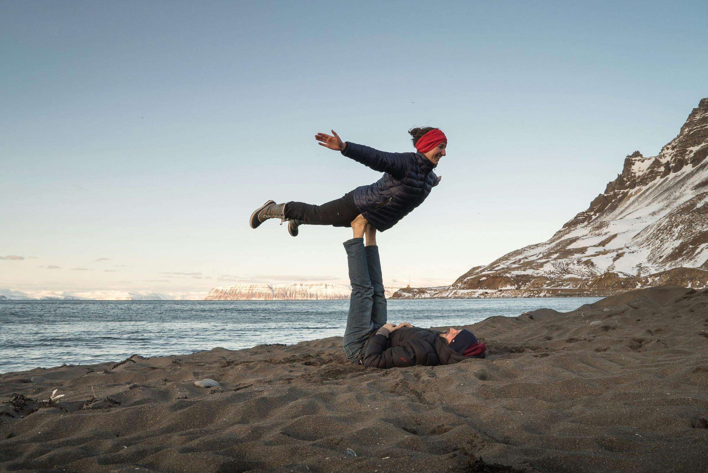 A couple does Yoga on a black sand beach. He is lying on his back and she is balancing on his feet. Mountains with snow in the background.