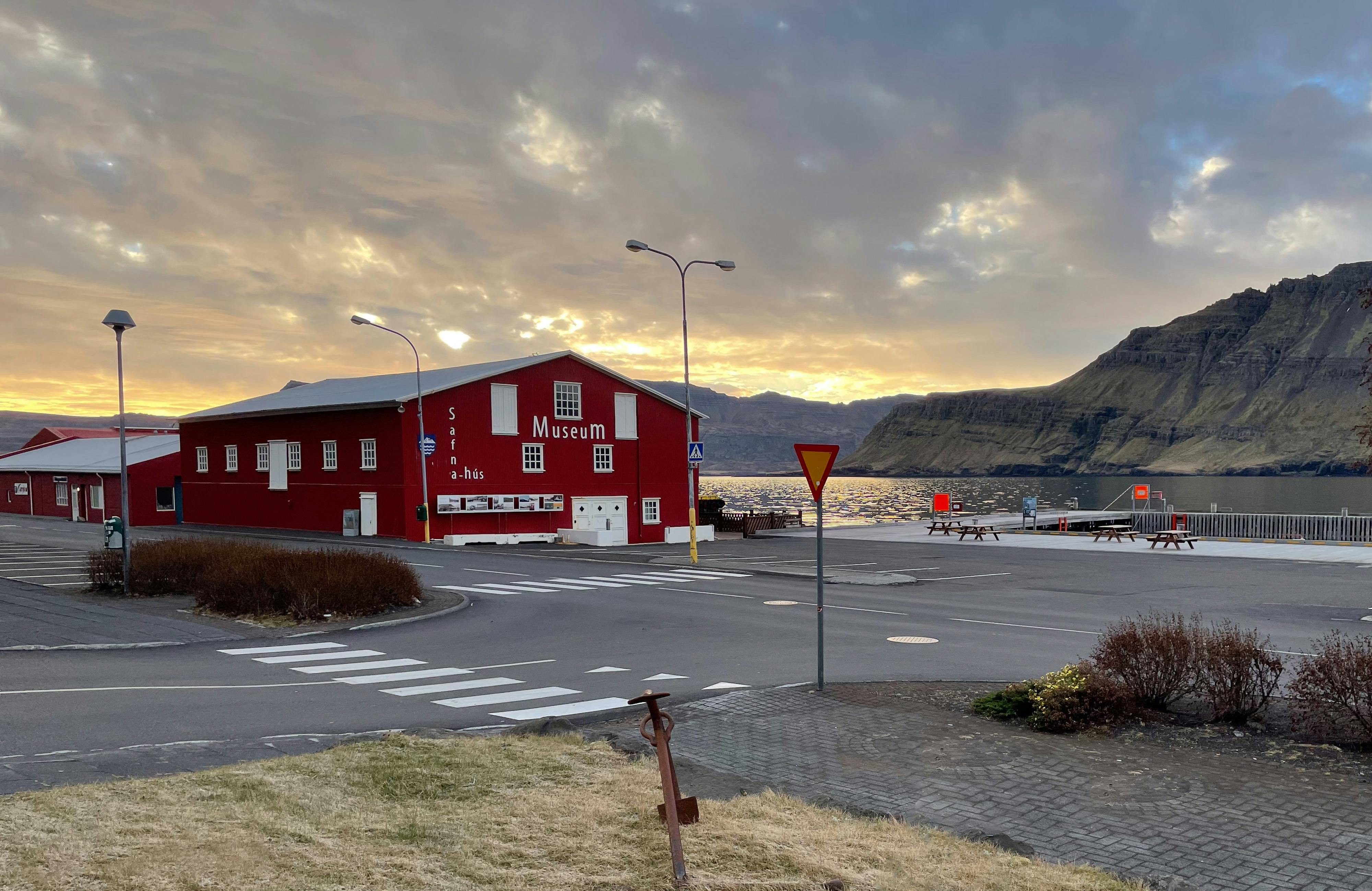 A wooden red museum building on the bank of a marina with tall mountains in the back