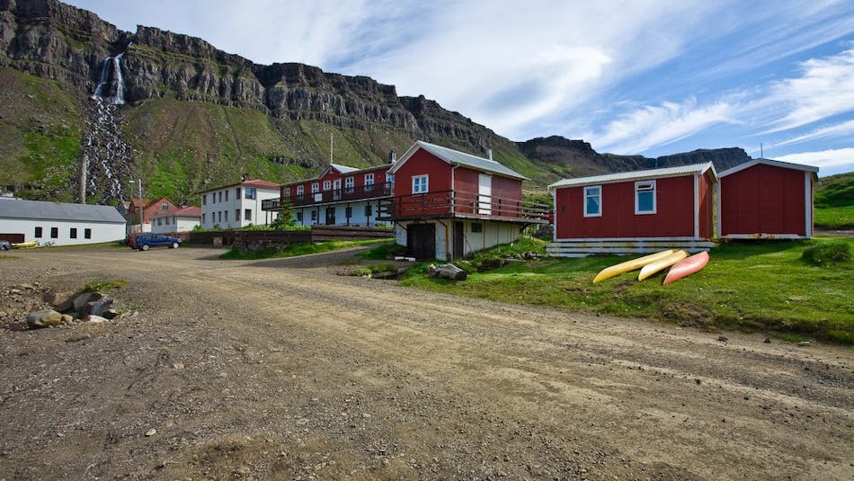 Red houses and the Hotel Djúpavik visible in the foreground, in the background there is the waterfall