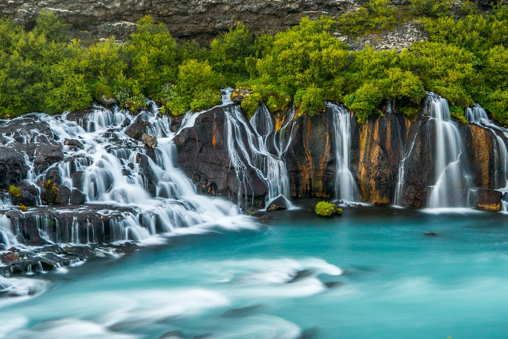 Series of spring waterfalls flowing from under a birch-grown lava into a white-blue colored glacier river
