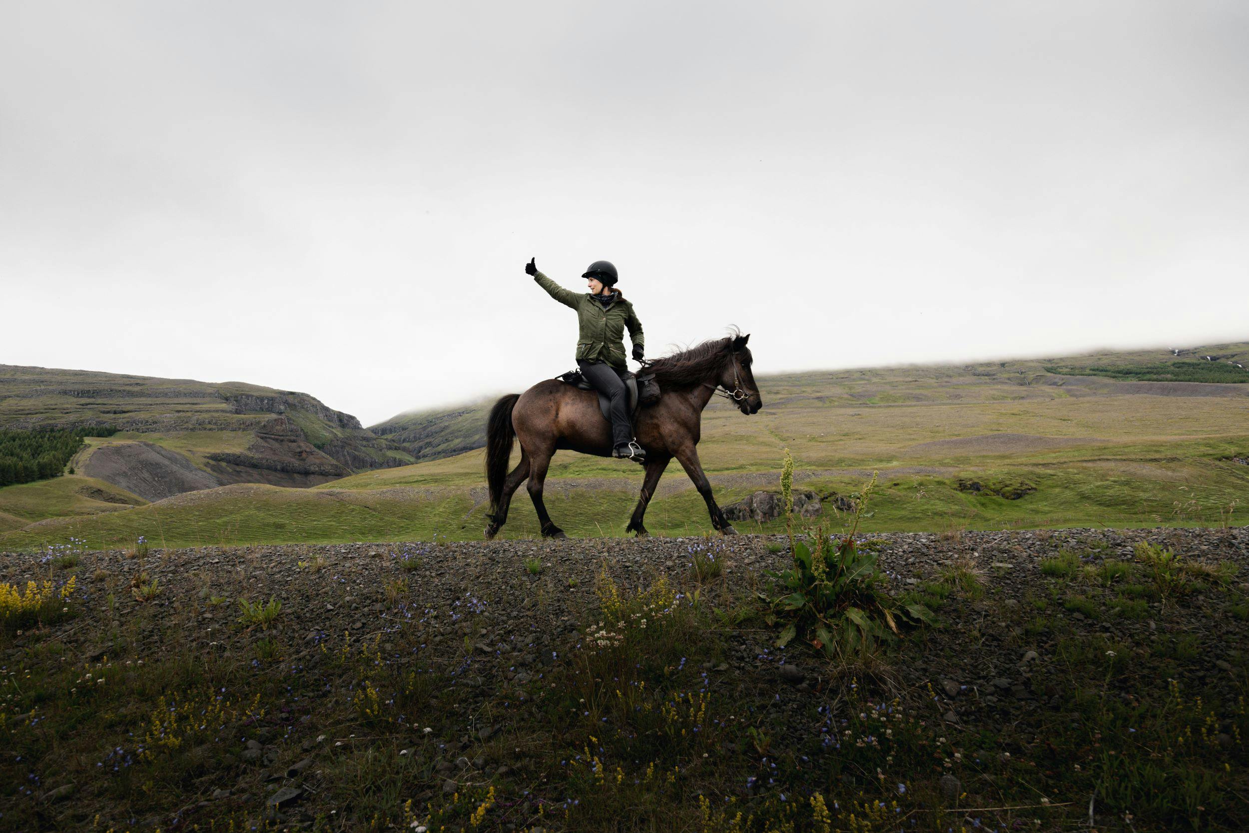 A woman riding a dark horse turns around, and gives a thumbs-up. to fellow riders.