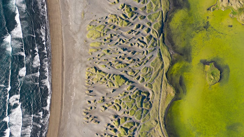 An aerial photo of a black sand beach, sand dunes and a green lake