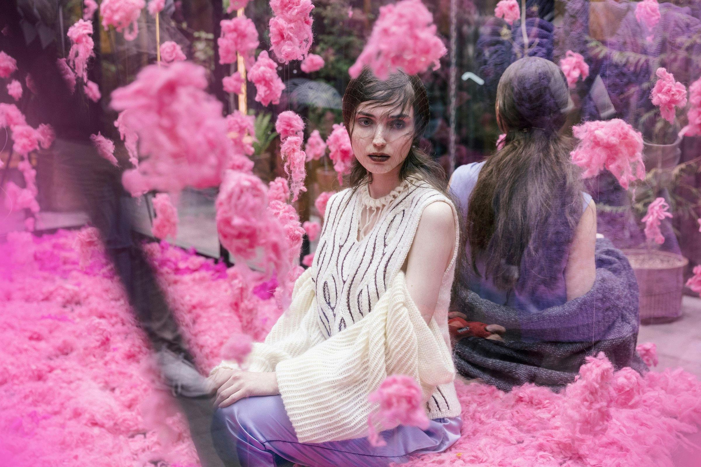 A woman sitting in pink coloured wool pieces draped on the floor and hanging from the ceiling. She is dressed in beige knitwear. 