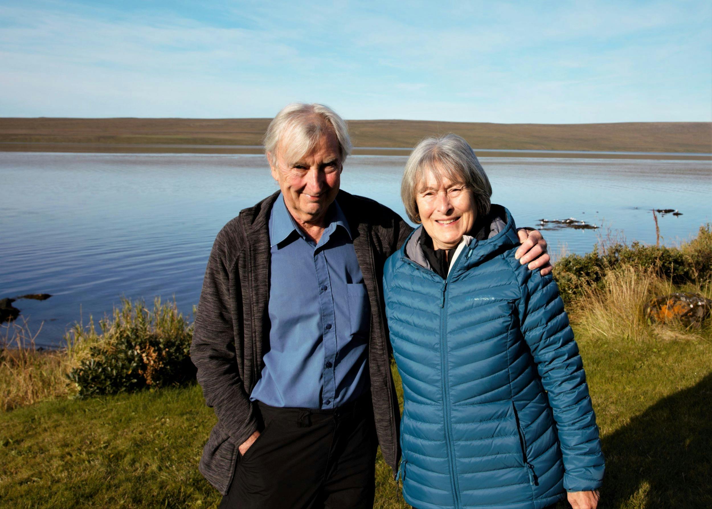 Elderly couple standing on a meadow, visible in the background is the ocean and some birds , the sun is shining