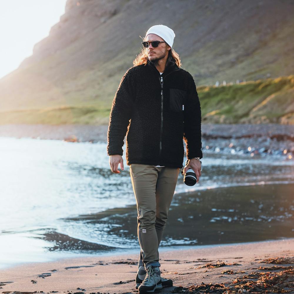 The Icelandic male model Rúrik Gíslason standing on a black beach during sundown, wearing hiking pants, a fleece jacket, white beanie and sunglasses. Mountains in the background