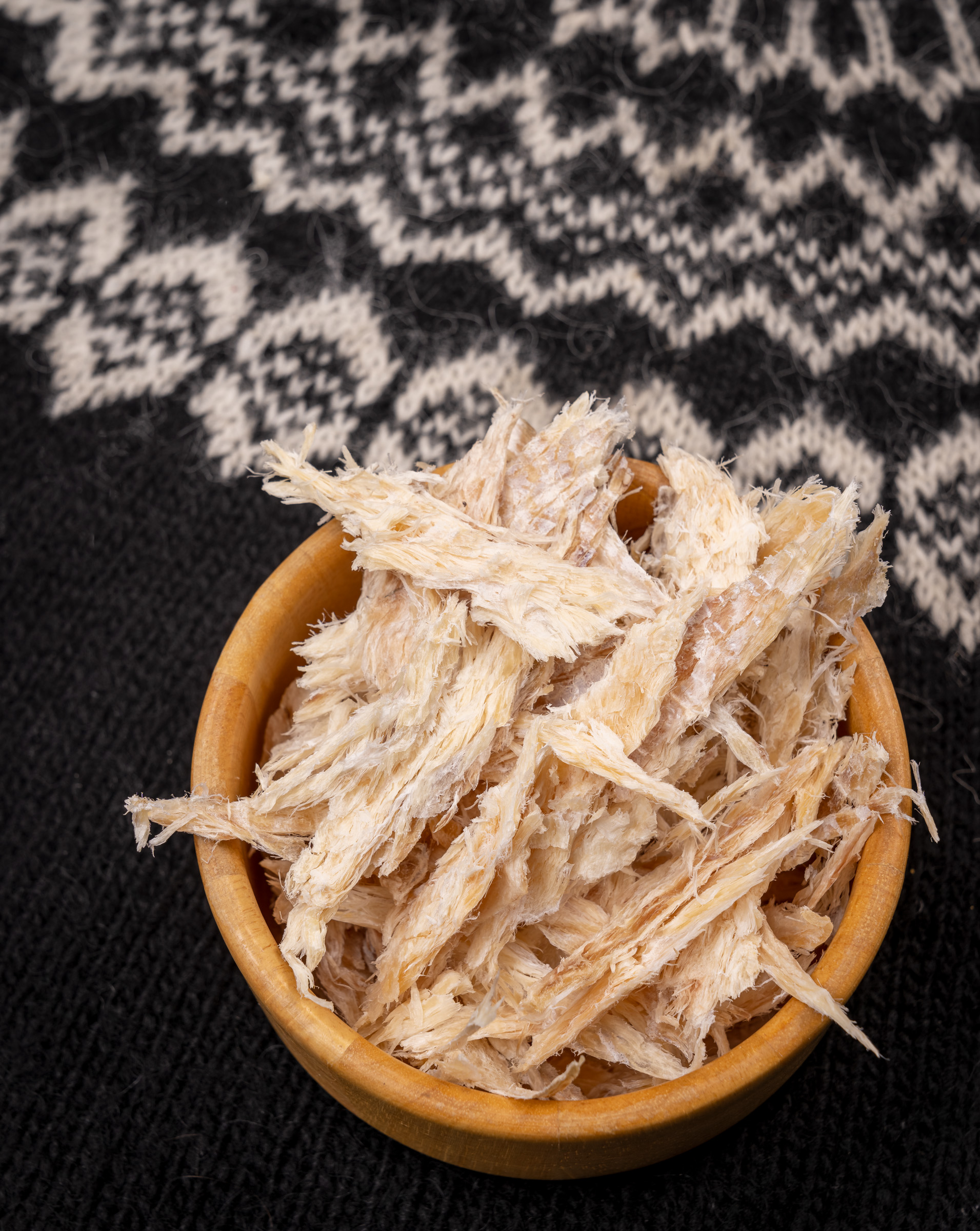 A wooden bowl filled with pieces of traditional Icelandic harðfiskur (dried fish), set against a background with an Icelandic wool sweater pattern