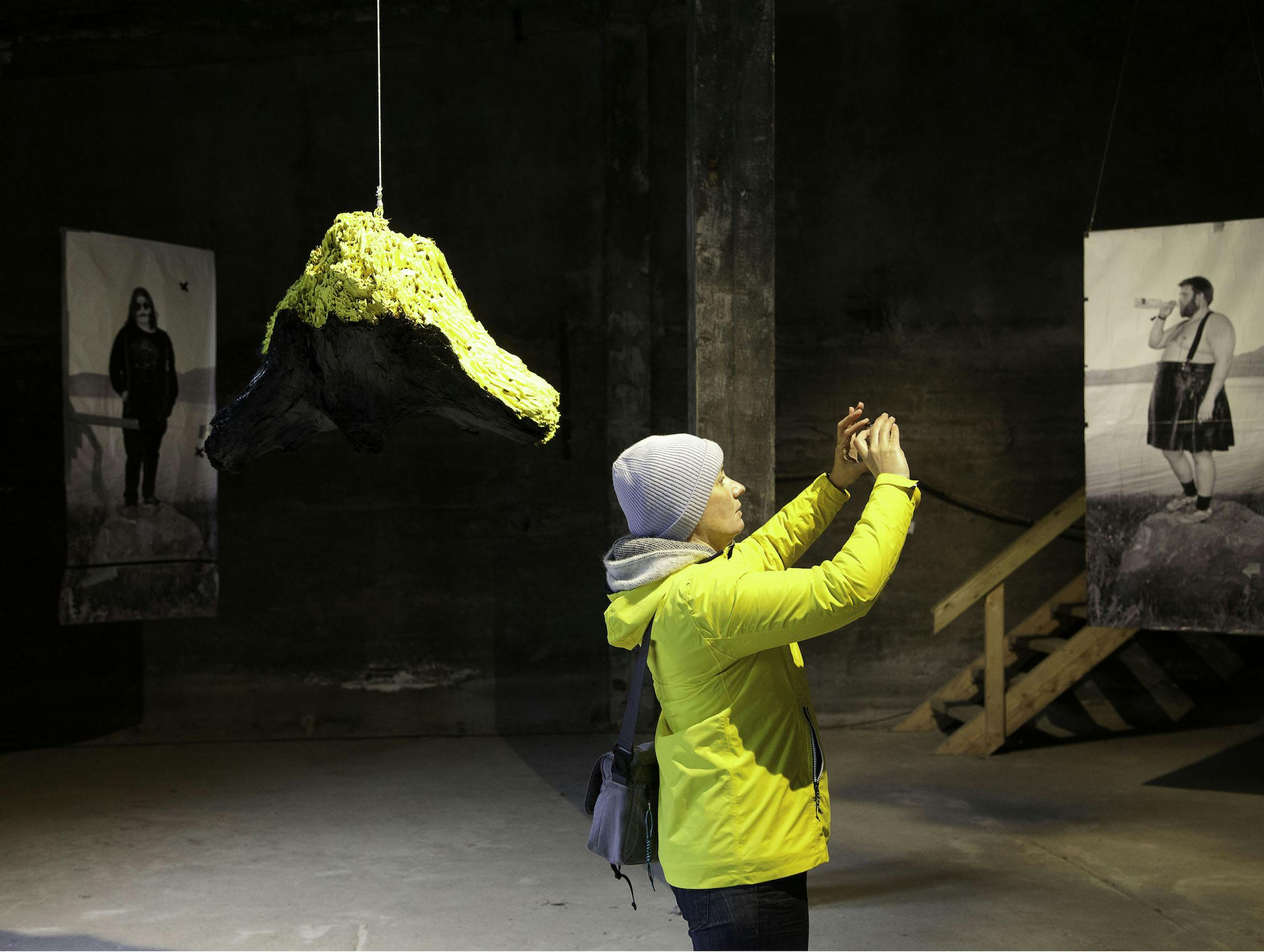 Visitor in yellow jacket photographing artworks of the exhibition in The Factory Art Exhibition in 2021. In the picture are visible the sculpture "I Am Calling For Colours" by  Emilie Dalum and photographs by  Attilio Solzi with the title 'MONOLITH'
