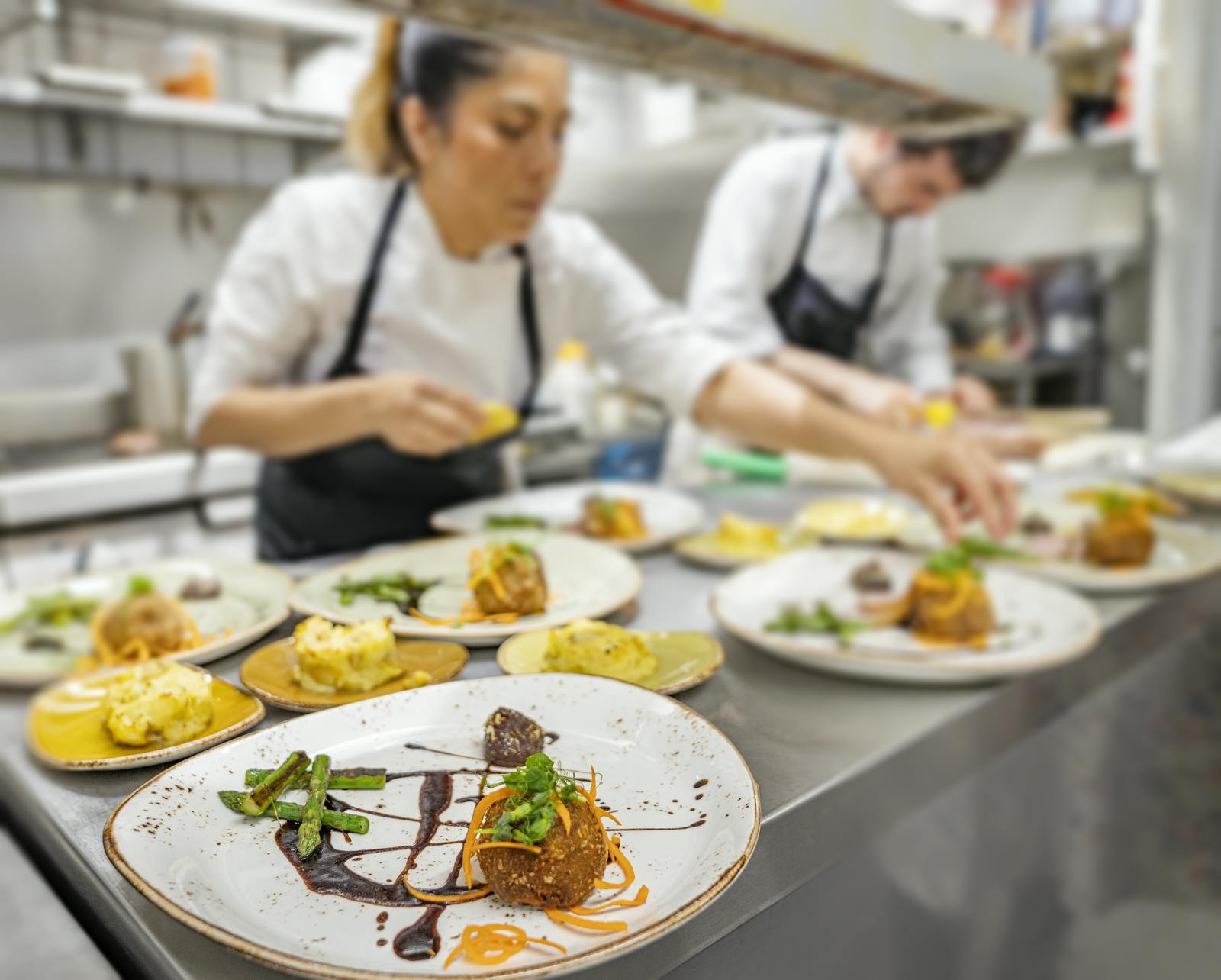 Two chefs arrange food in a restaurant kitchen, round platters with vegetables and sauce in front of them