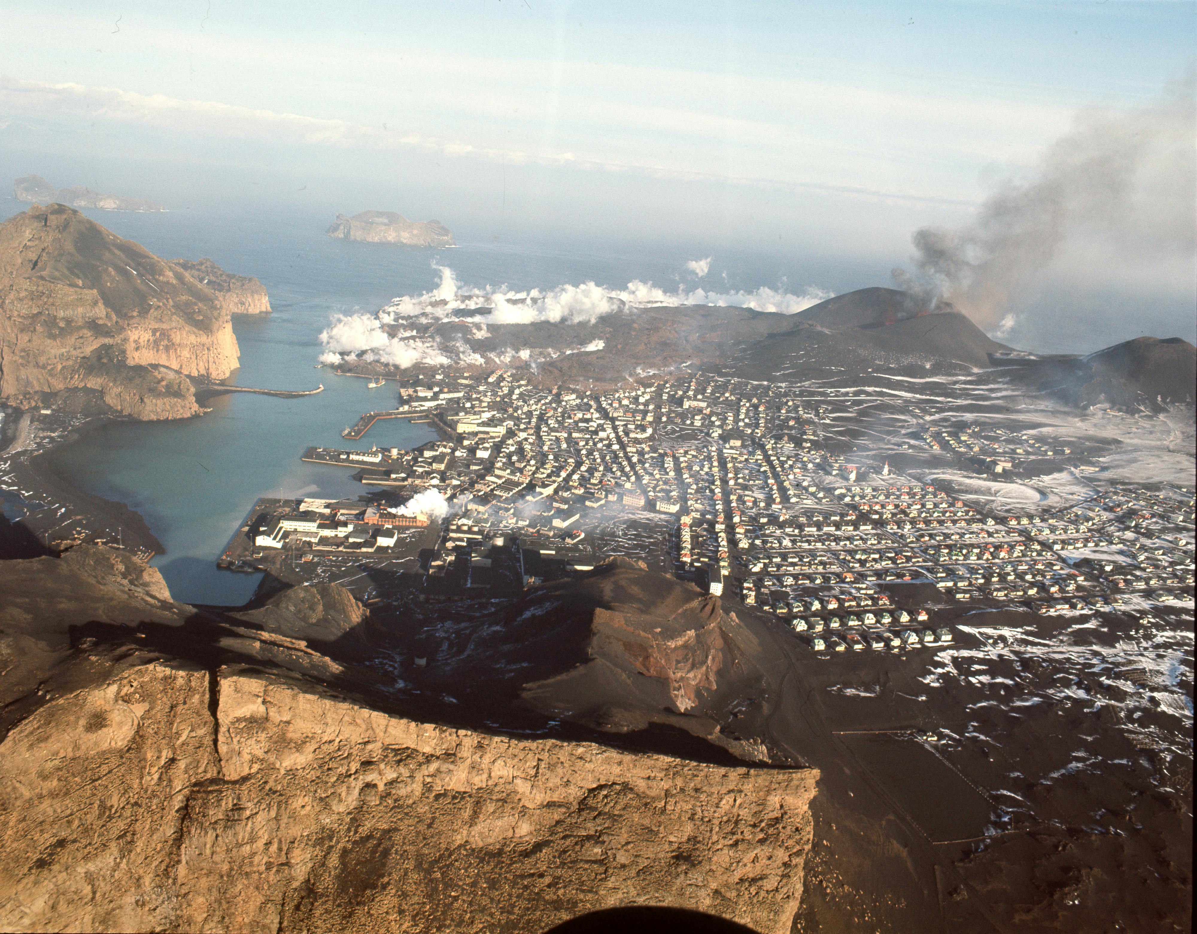 An erupting volcano spouting ash over a town on a small island