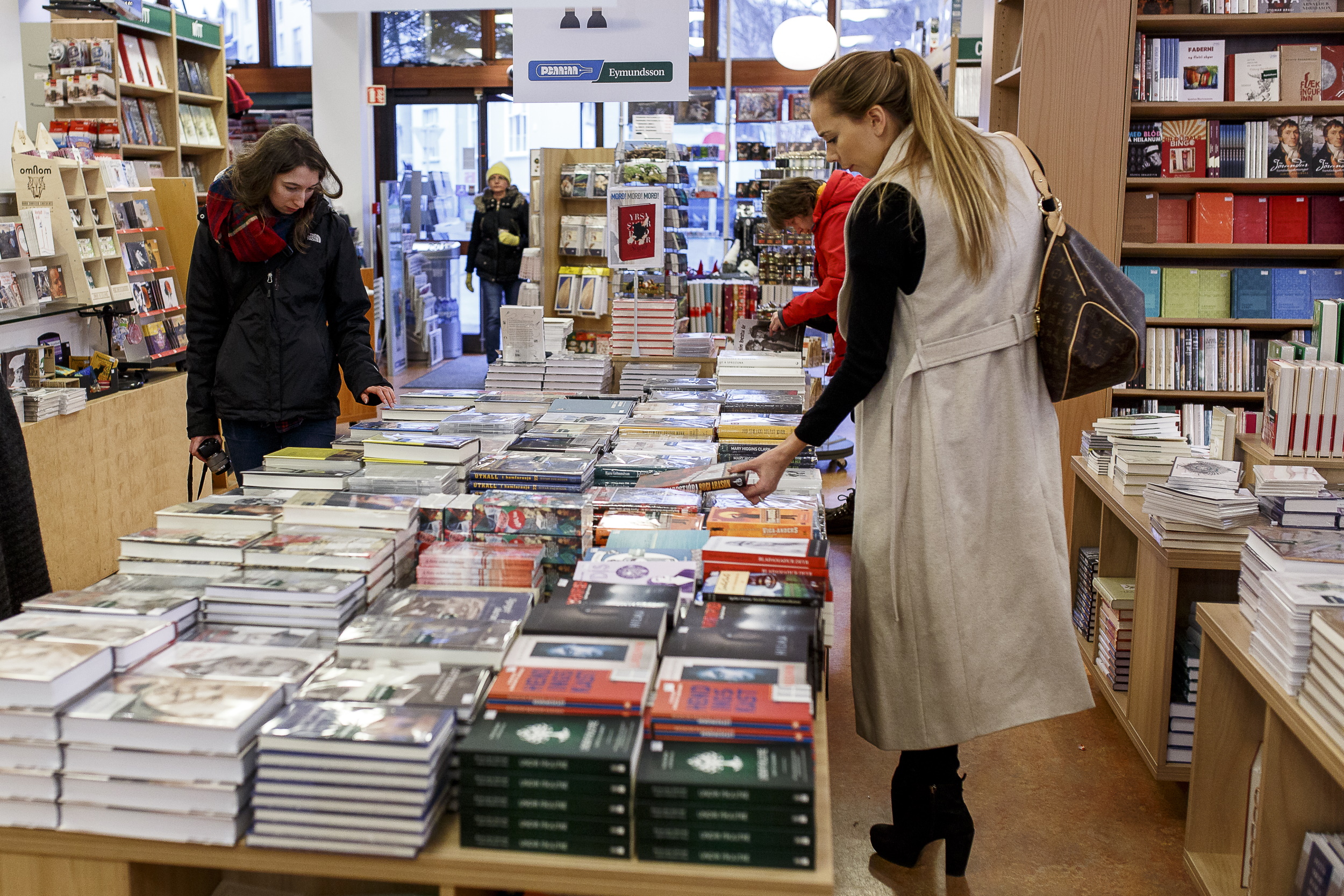 A woman browses stacks of new book titles displayed on large tables inside an Eymundsson bookstore. Shelves filled with books line the bright, inviting space, and other customers explore the aisles in the background. Eymundsson is an established bookstore chain found across Iceland.