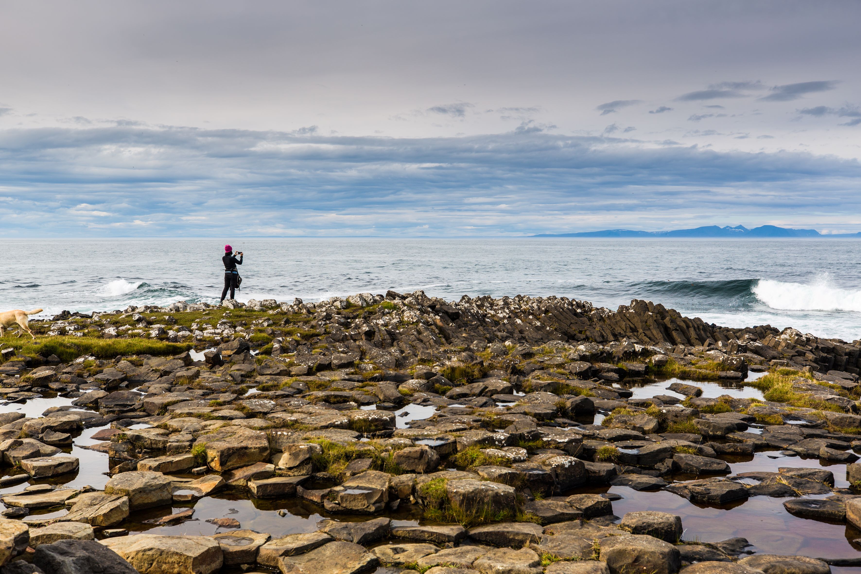 Woman standing on flat basalt stones looking towards the ocean and taking pictures