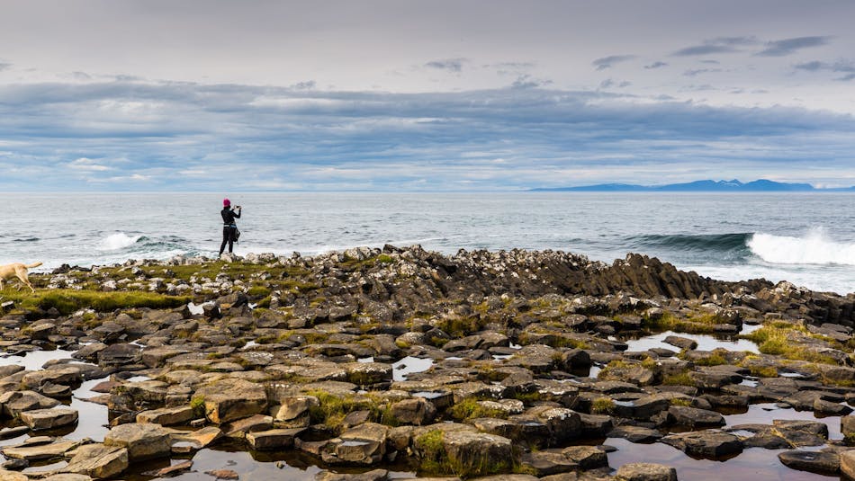 Woman standing on flat basalt stones looking towards the ocean and taking pictures