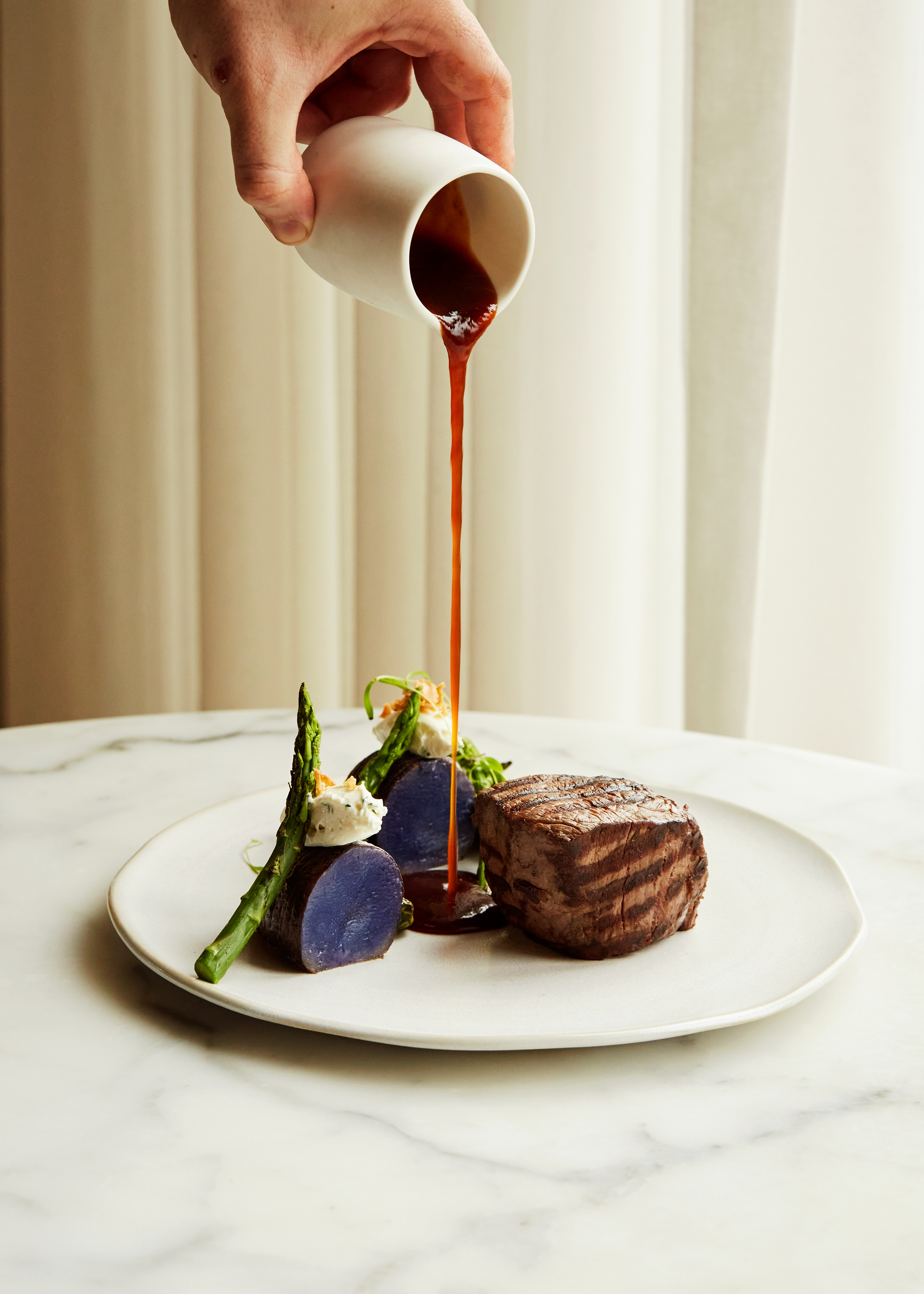 A rich sauce is poured over a beautifully plated dish featuring grilled meat, purple potatoes, and green asparagus on a white plate at Reykjavík Edition. 