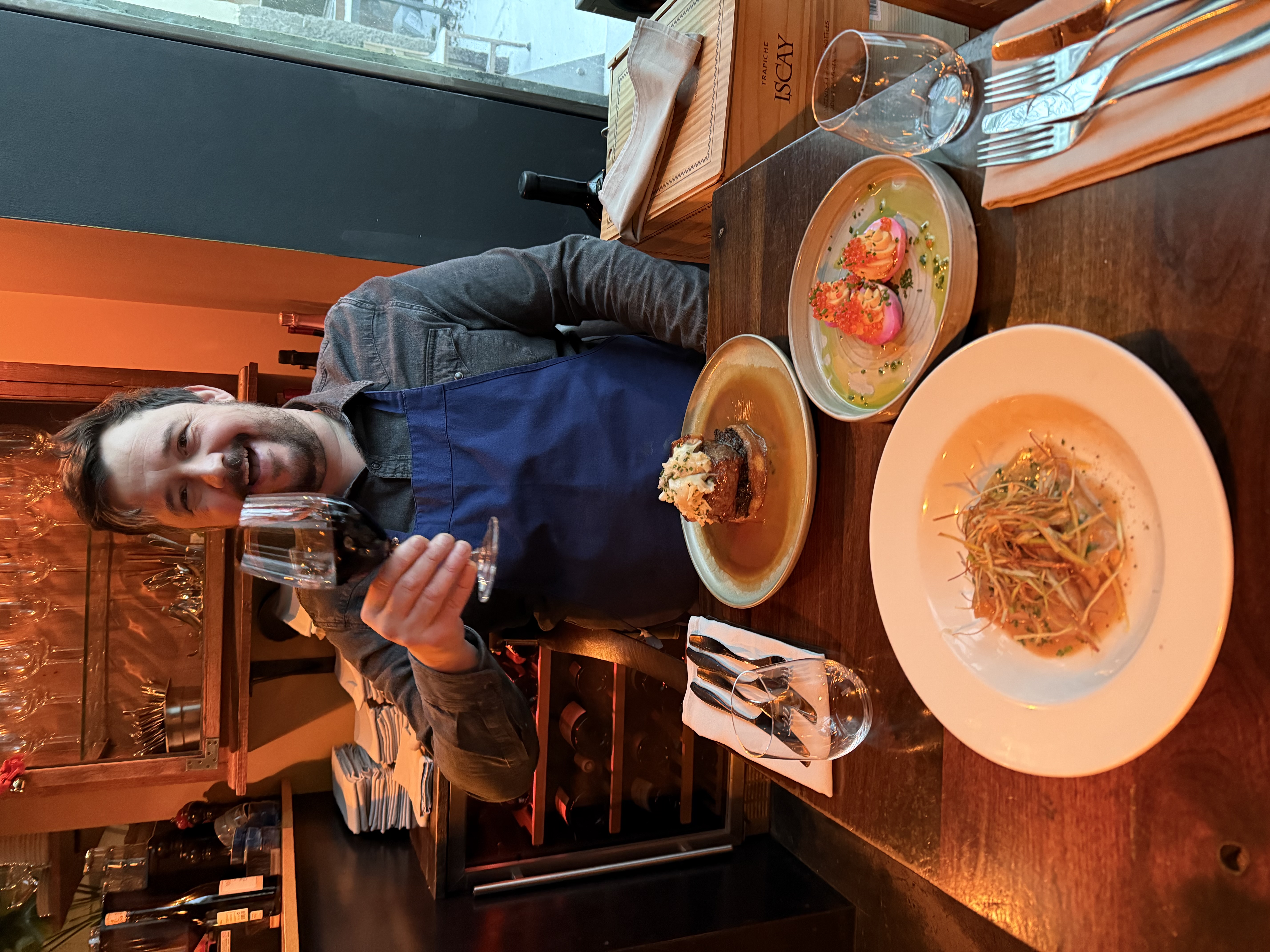 A guest raises a glass of red wine and smiles at Hosiló restaurant in Reykjavík, seated at a table with three beautifully plated dishes.