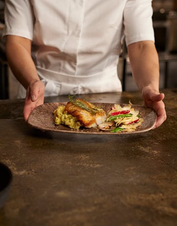 Chef serving a plated Icelandic cod dish with vegetables and garnish.