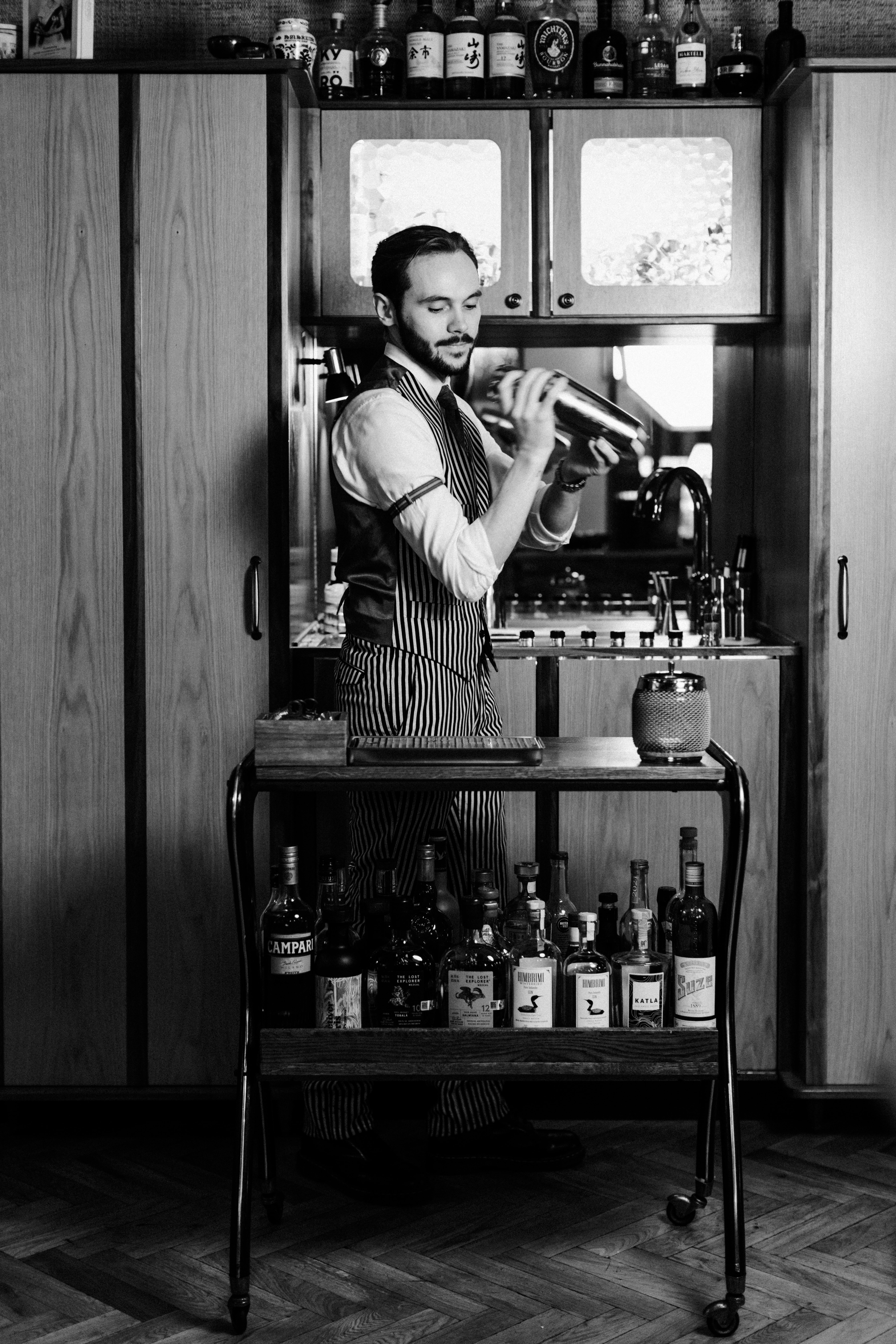 Black-and-white photo of a bartender at ÓX restaurant, wearing a striped vest and shaking a cocktail behind a cart stocked with bottles and bar tools
