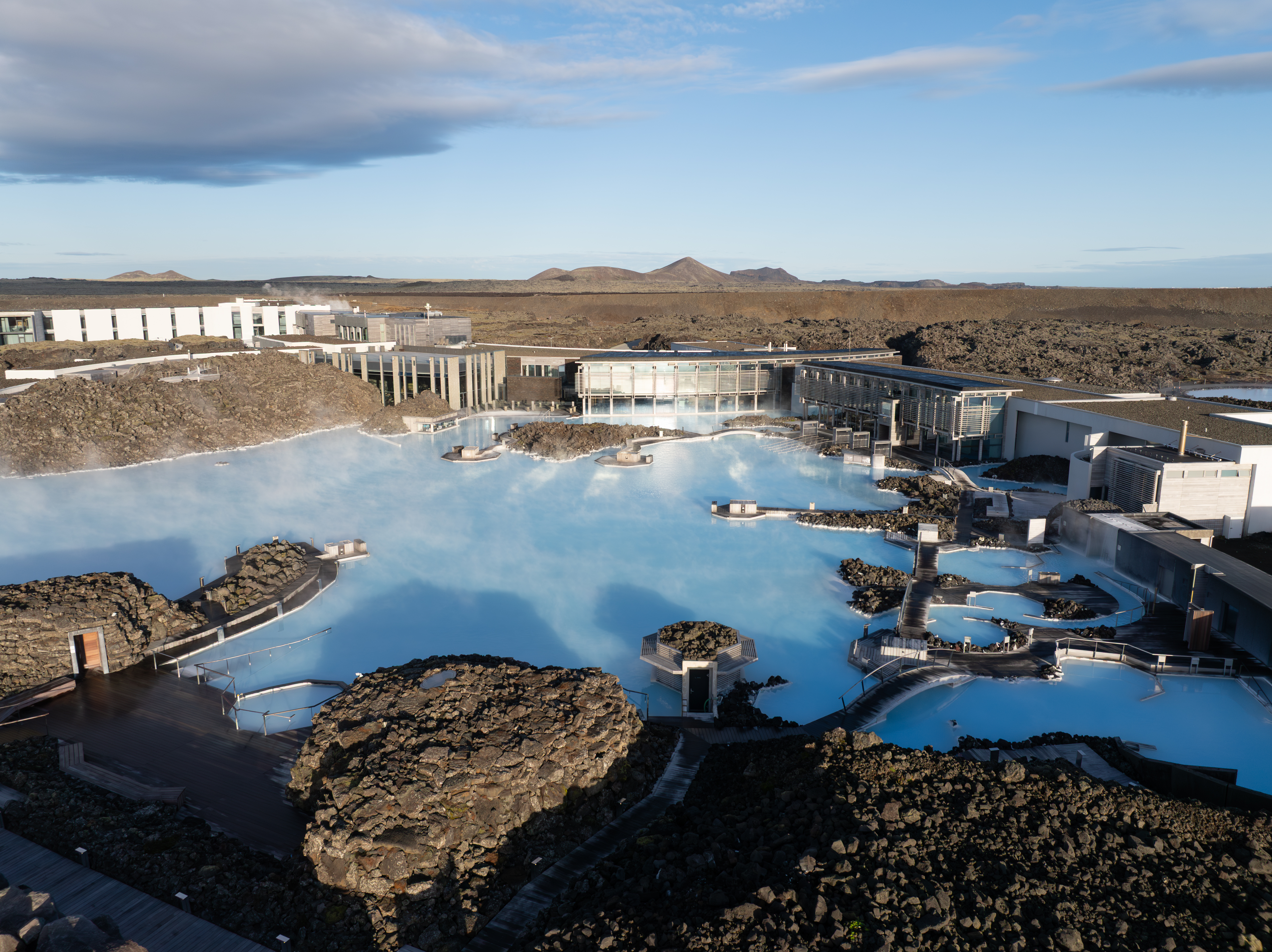 A wide, elevated view of the Blue Lagoon in Iceland, showing its milky blue geothermal waters surrounded by dark volcanic lava fields and modern spa buildings under a clear sky.
