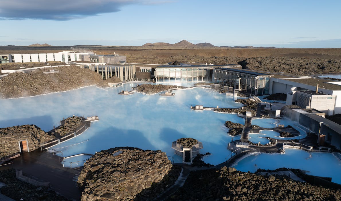 A wide, elevated view of the Blue Lagoon in Iceland, showing its milky blue geothermal waters surrounded by dark volcanic lava fields and modern spa buildings under a clear sky.