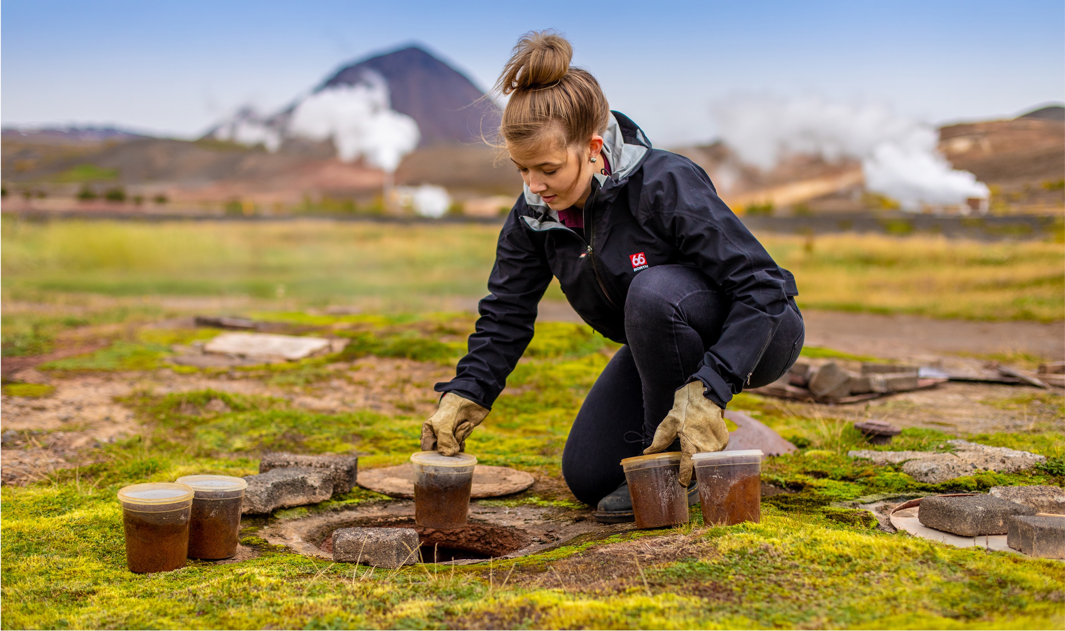 A woman baking rye bread in the ground near Mývatn with mountains and smoke in the background