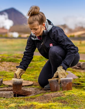 A woman baking rye bread in the ground near Mývatn with mountains and smoke in the background