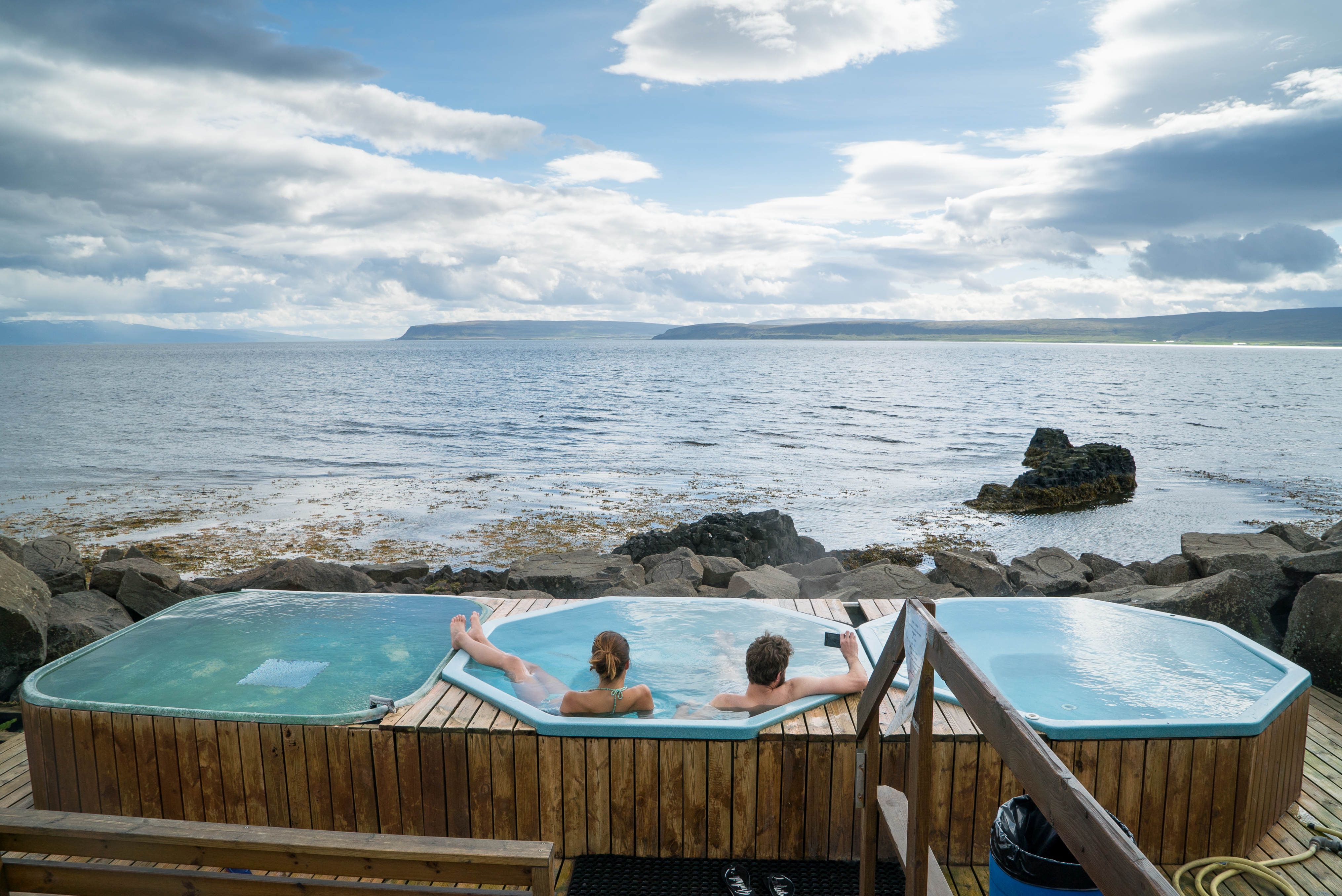 Two people relax in small outdoor hot pots built from wood on the rocky shoreline in Drangsnes. The tubs overlook a calm sea with distant cliffs and cloud-filled skies stretching across the horizon.