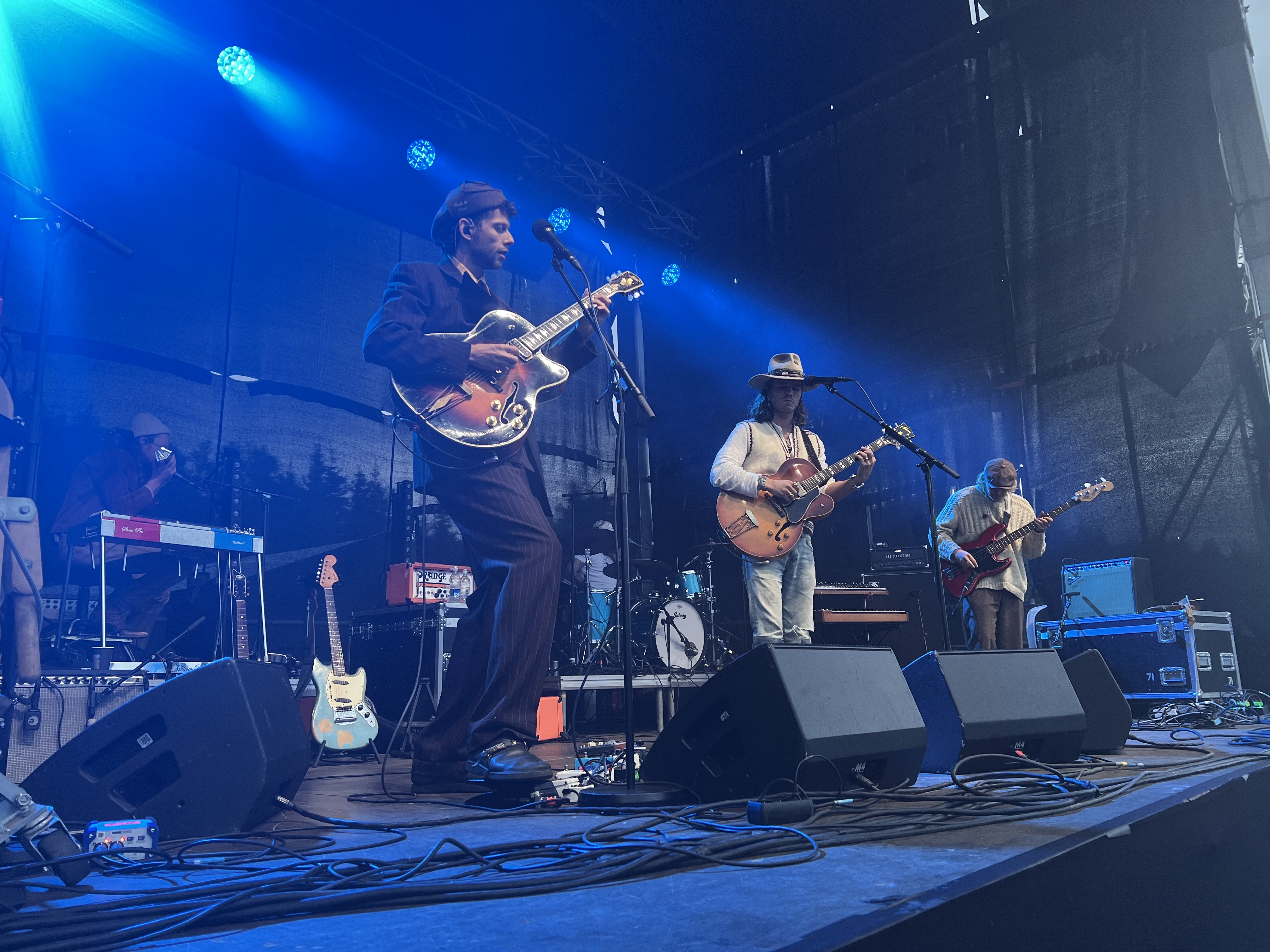 The Icelandic band Kaleo performs live on an outdoor stage under blue lighting, with two guitarists at microphones and other band members and instruments visible behind them.
