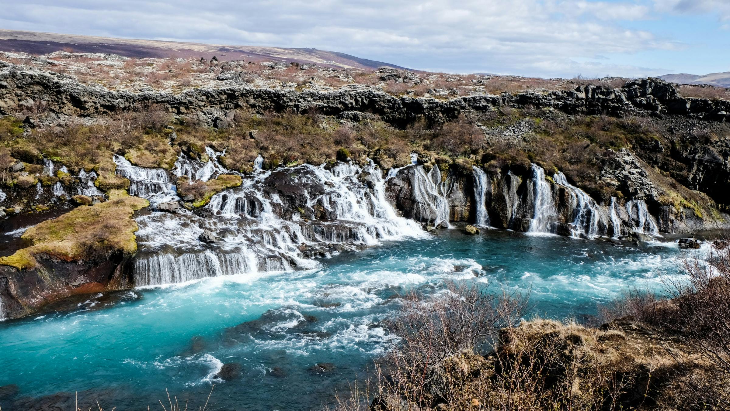 Spring waterfalls flowing from under a lava into the white-blue colored glacier river Hvita