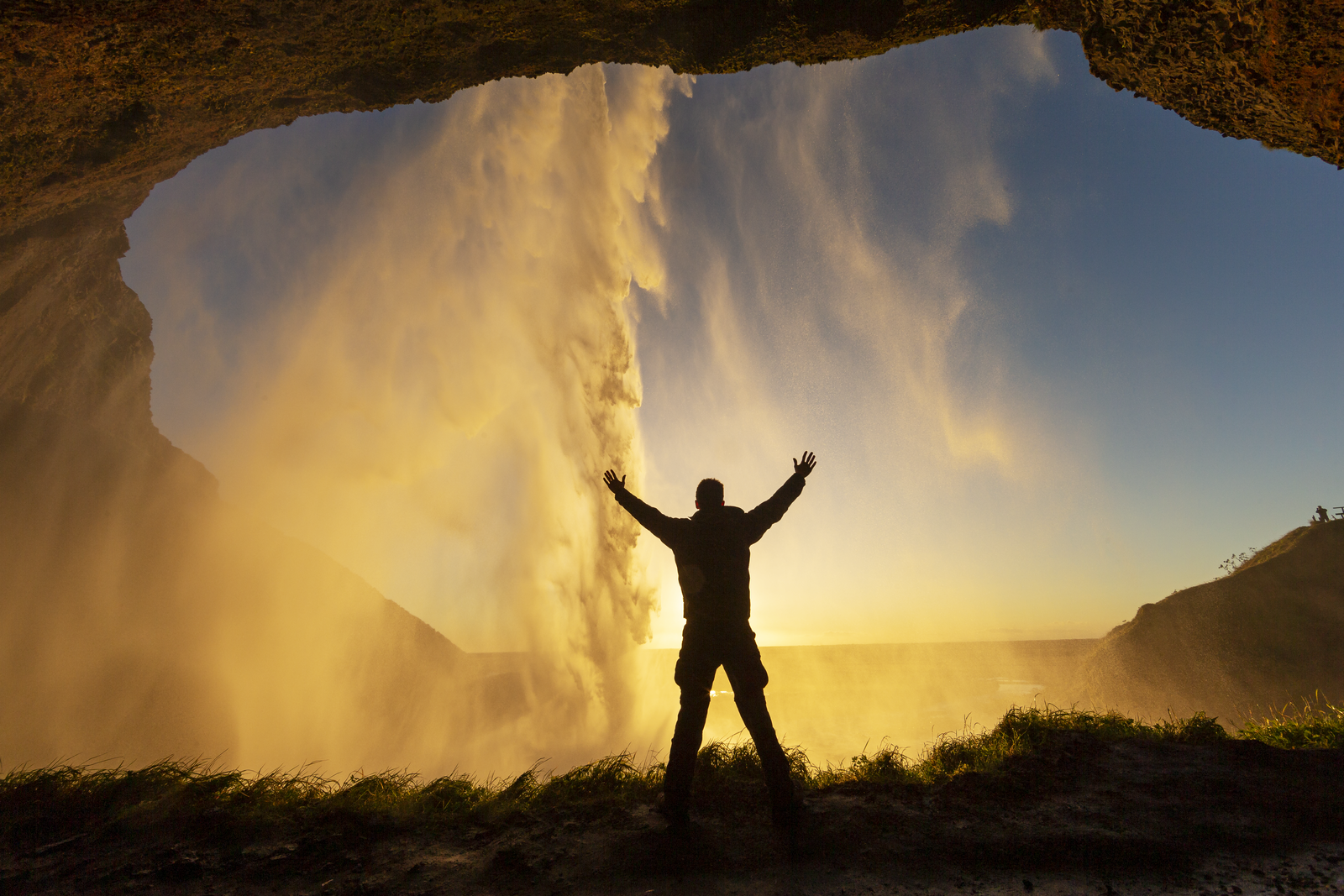 Person standing behind Seljalandsfoss waterfall in Iceland at sunset, silhouetted with arms raised as water cascades down in golden light.