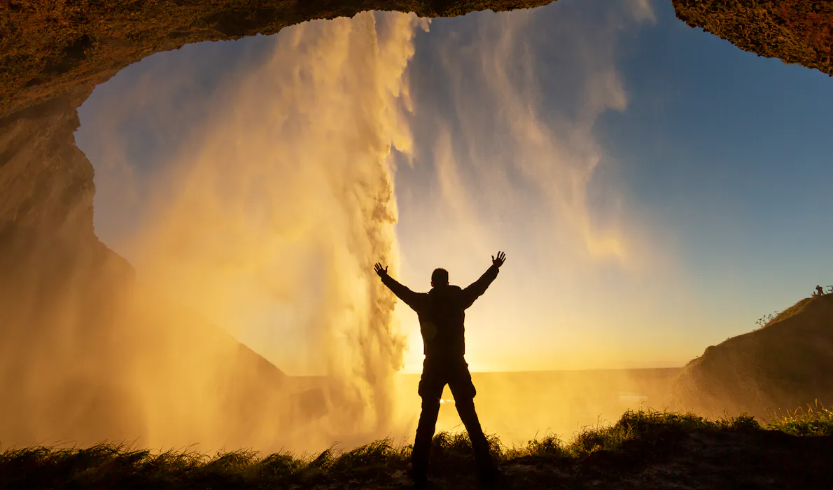 Person standing behind Seljalandsfoss waterfall in Iceland at sunset, silhouetted with arms raised as water cascades down in golden light.