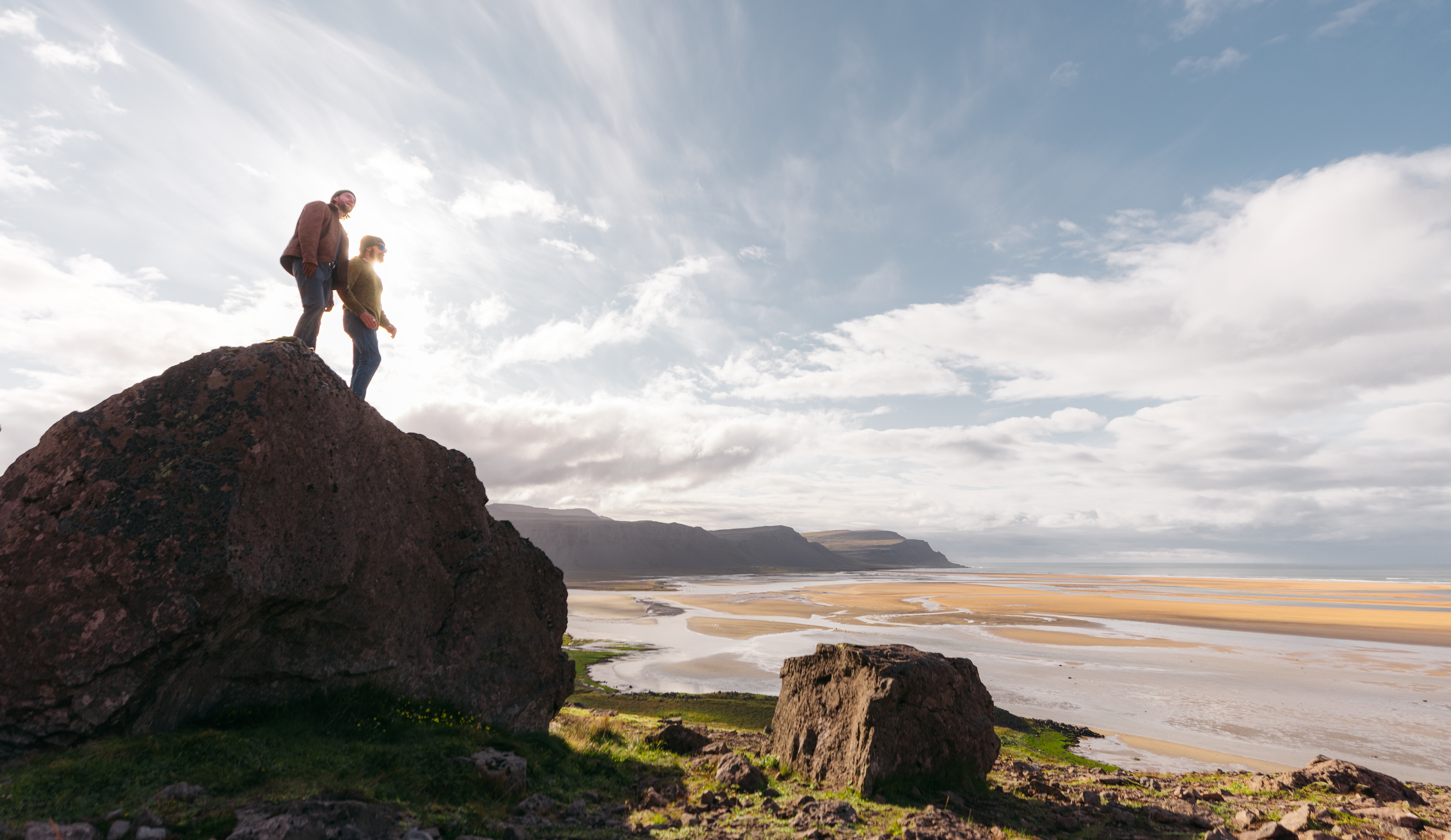 Two people standing on a rocky cliff overlooking Rauðasandur beach in the Westfjords of Iceland under a bright summer sky.