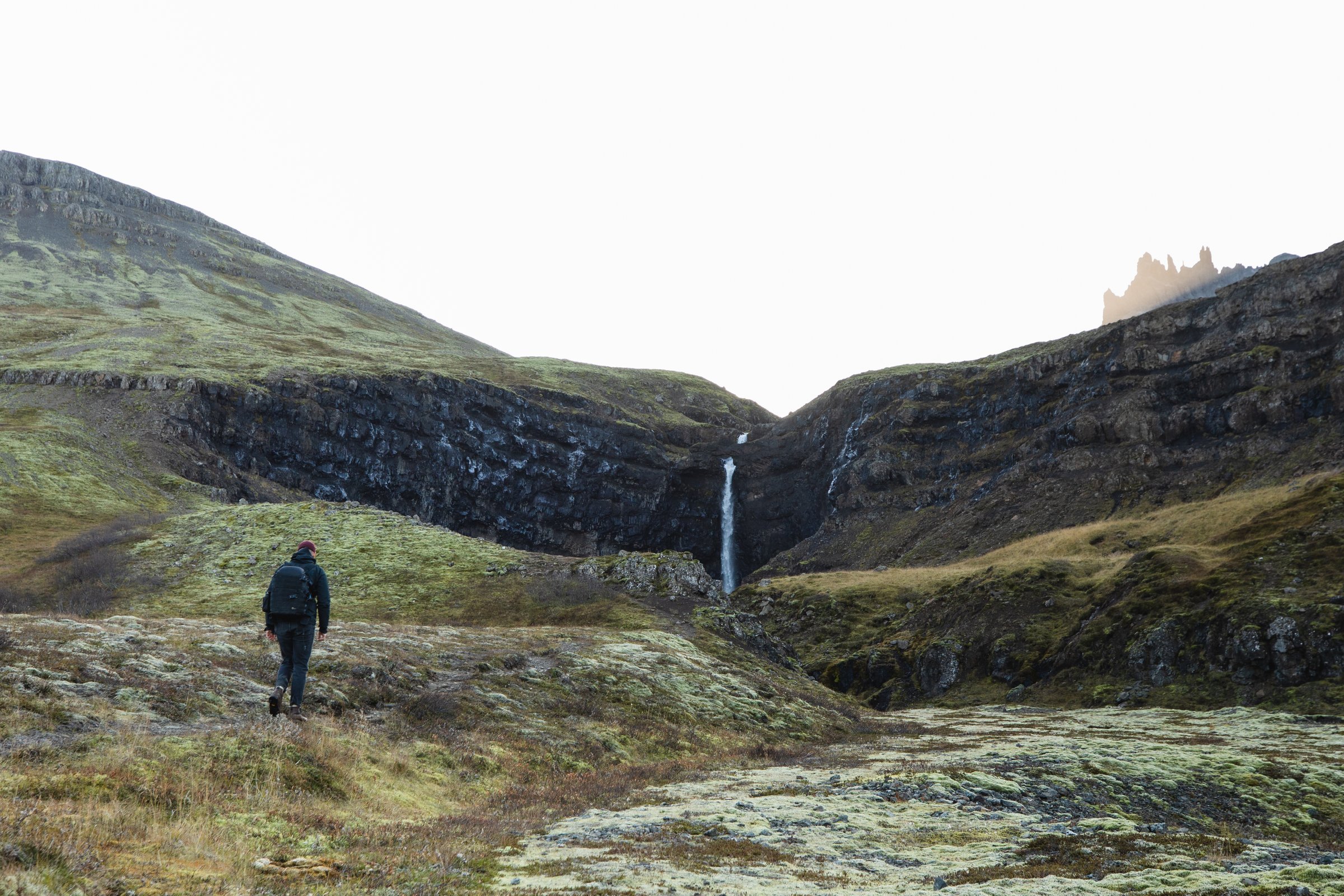 Hiker walking through a moss-covered valley toward Flögufoss waterfall in East Iceland, surrounded by rugged hills and cliffs.