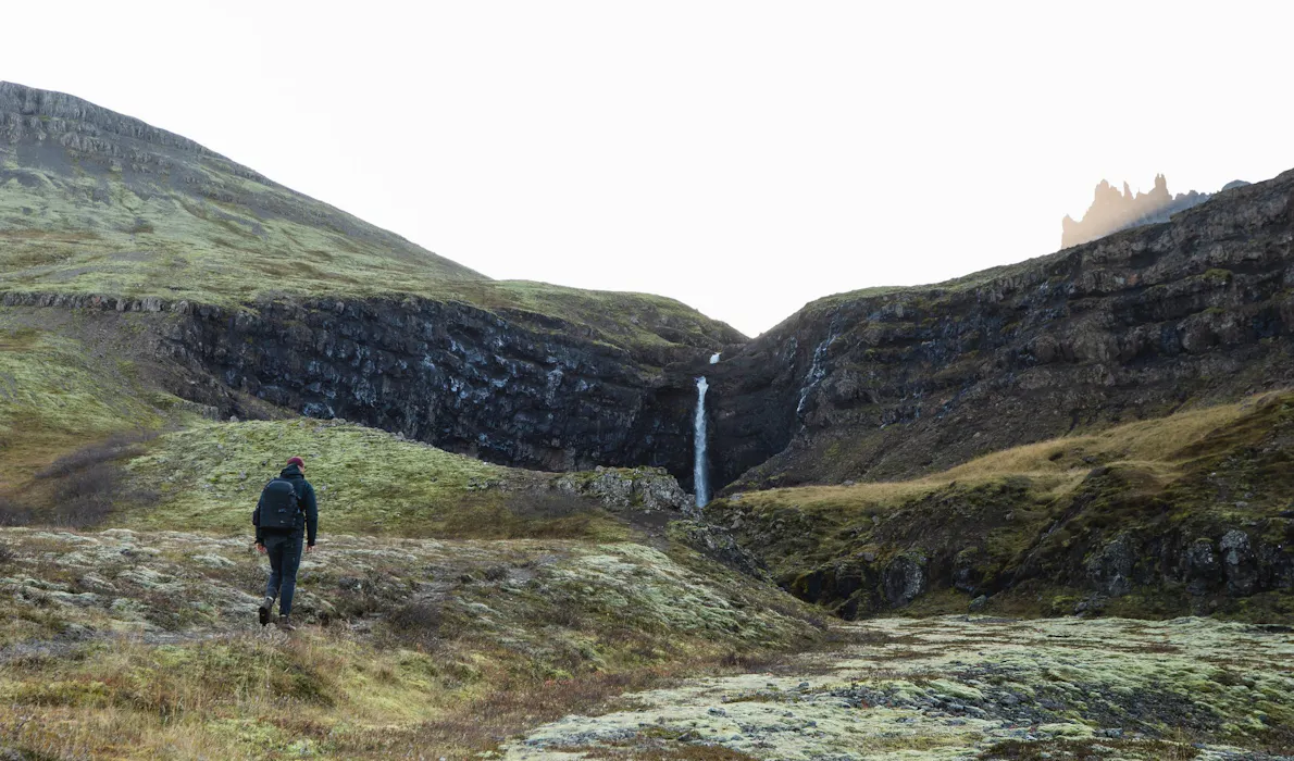 Hiker walking through a moss-covered valley toward Flögufoss waterfall in East Iceland, surrounded by rugged hills and cliffs.