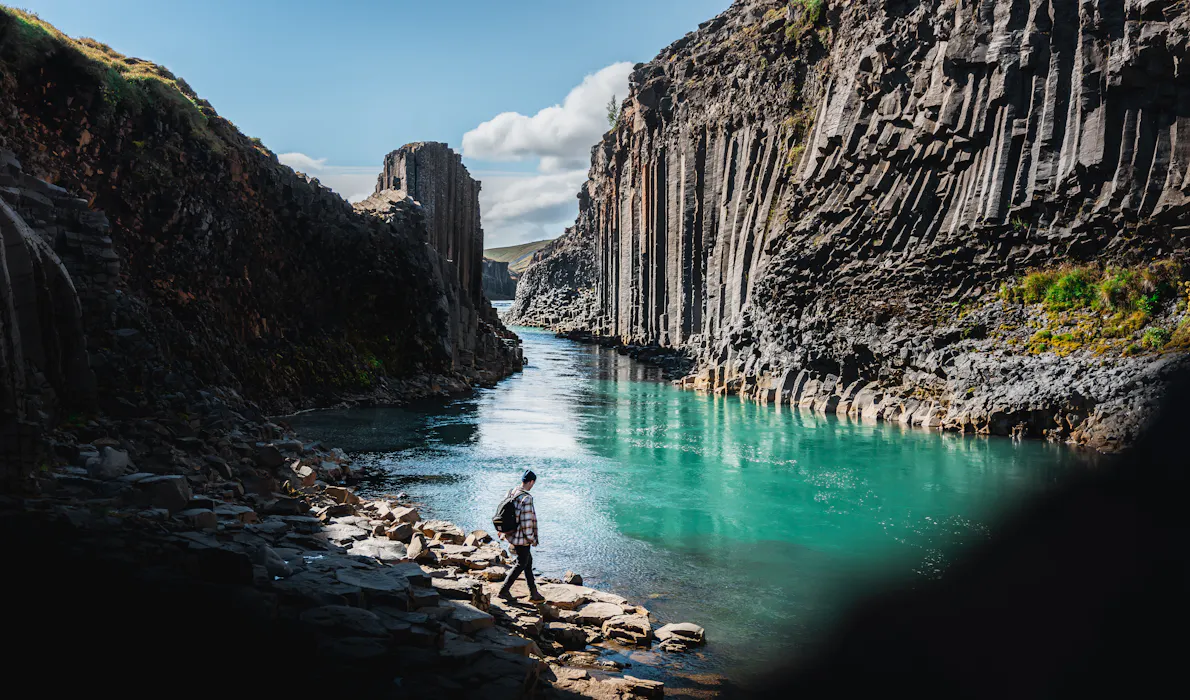 A person walking by Stuðlagil Canyon, with turquoise glacial waters flowing between steep basalt cliffs in East Iceland.