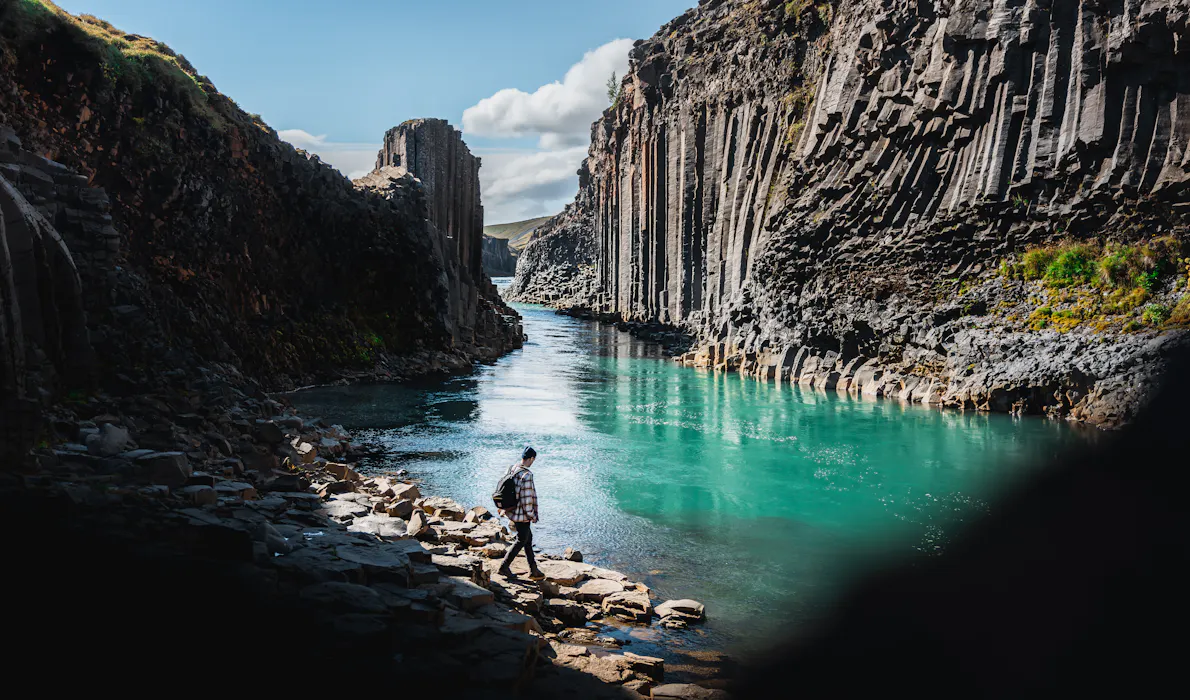 A person walking by Stuðlagil Canyon, with turquoise glacial waters flowing between steep basalt cliffs in East Iceland.