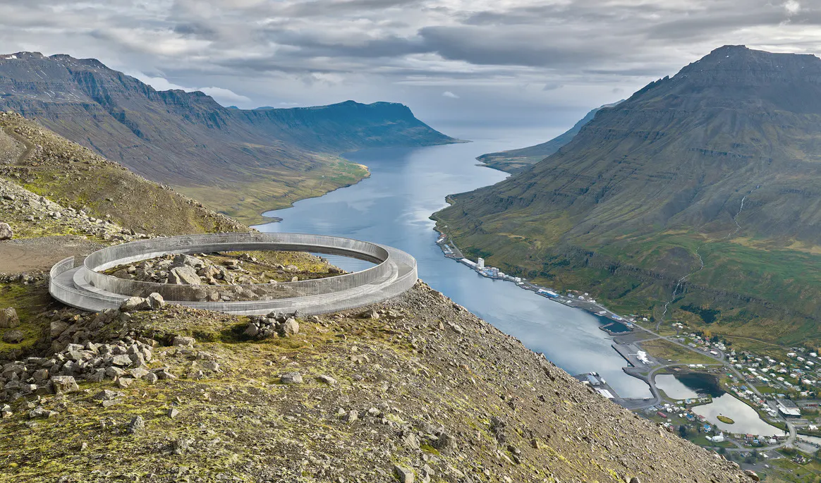 A circular viewing platform overlooks Seyðisfjörður from a mountainside high above the fjord. Steep mountains rise on either side of the water, with patches of snow, drifting mist, and rugged volcanic terrain creating a dramatic East Iceland landscape.
